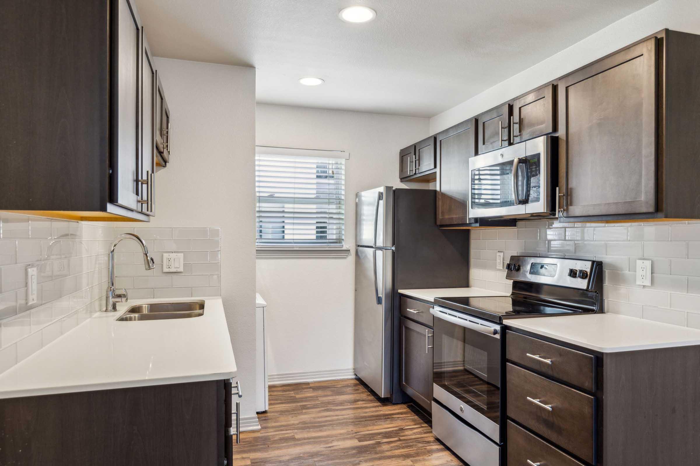 Modern kitchen featuring dark wood cabinetry, stainless steel appliances, and white countertops. Includes a sink, stove, oven, and refrigerator. The space has a bright atmosphere with overhead lighting and a window with blinds, enhancing the overall appeal.