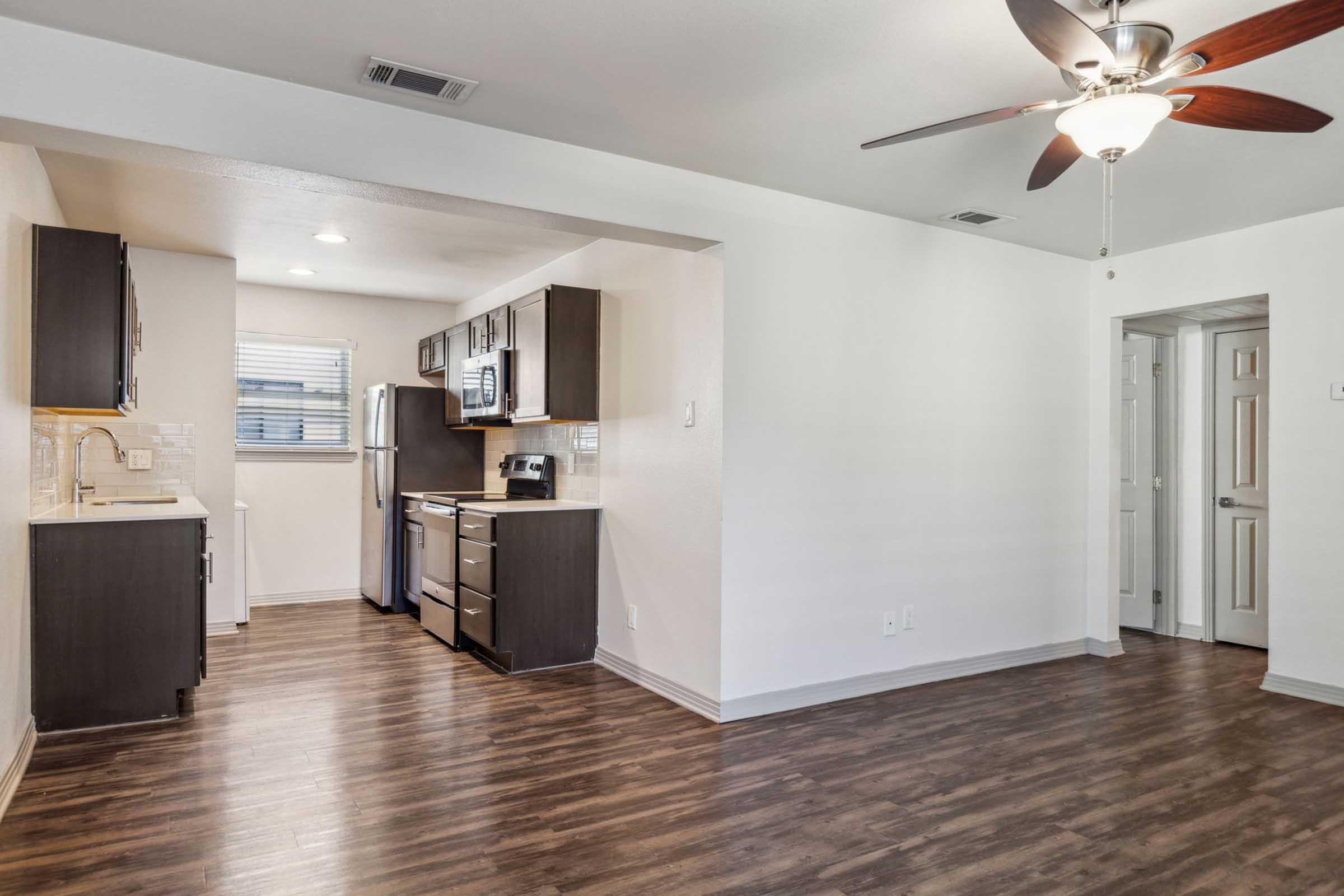 A modern kitchen and living area featuring dark wood cabinets, stainless steel appliances, and a ceiling fan. The space has a light-colored wall and laminate flooring, with a window providing natural light. A doorway leads to another room in the background.