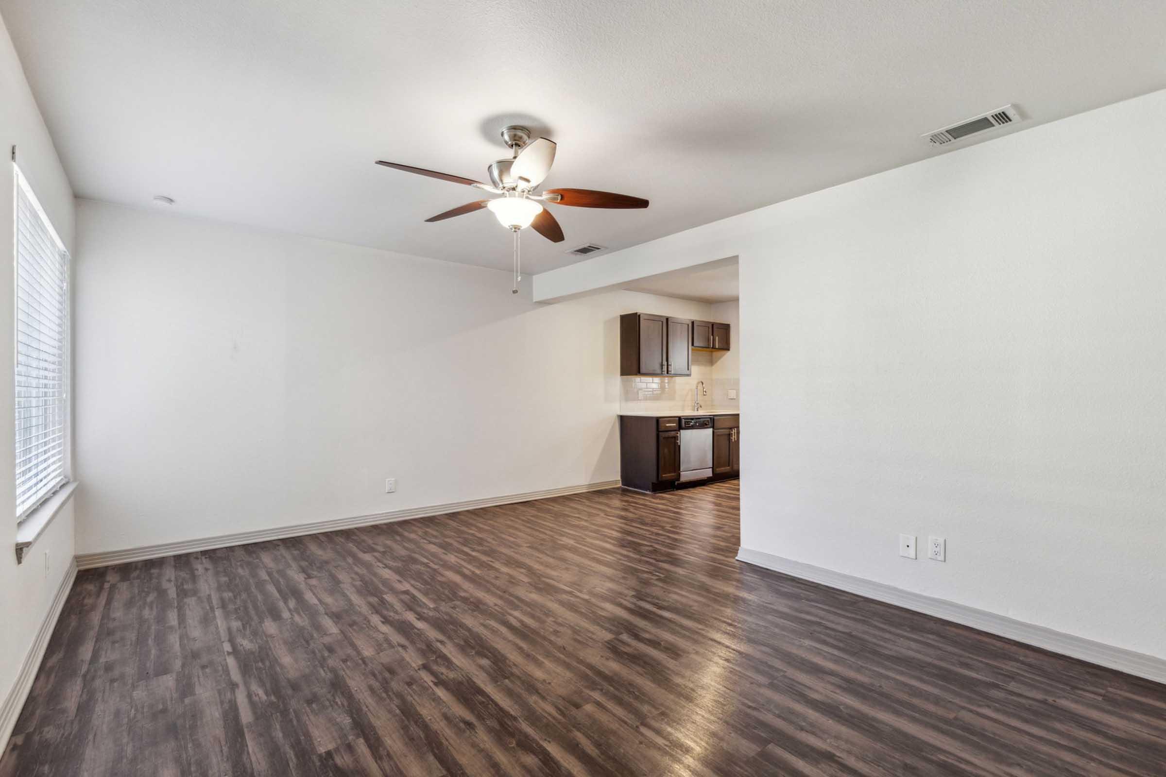 Spacious living area with a ceiling fan and large window providing natural light. The room features dark laminate flooring and connects to a modern kitchen with dark cabinetry and stainless steel appliances. White walls create a bright and open atmosphere.