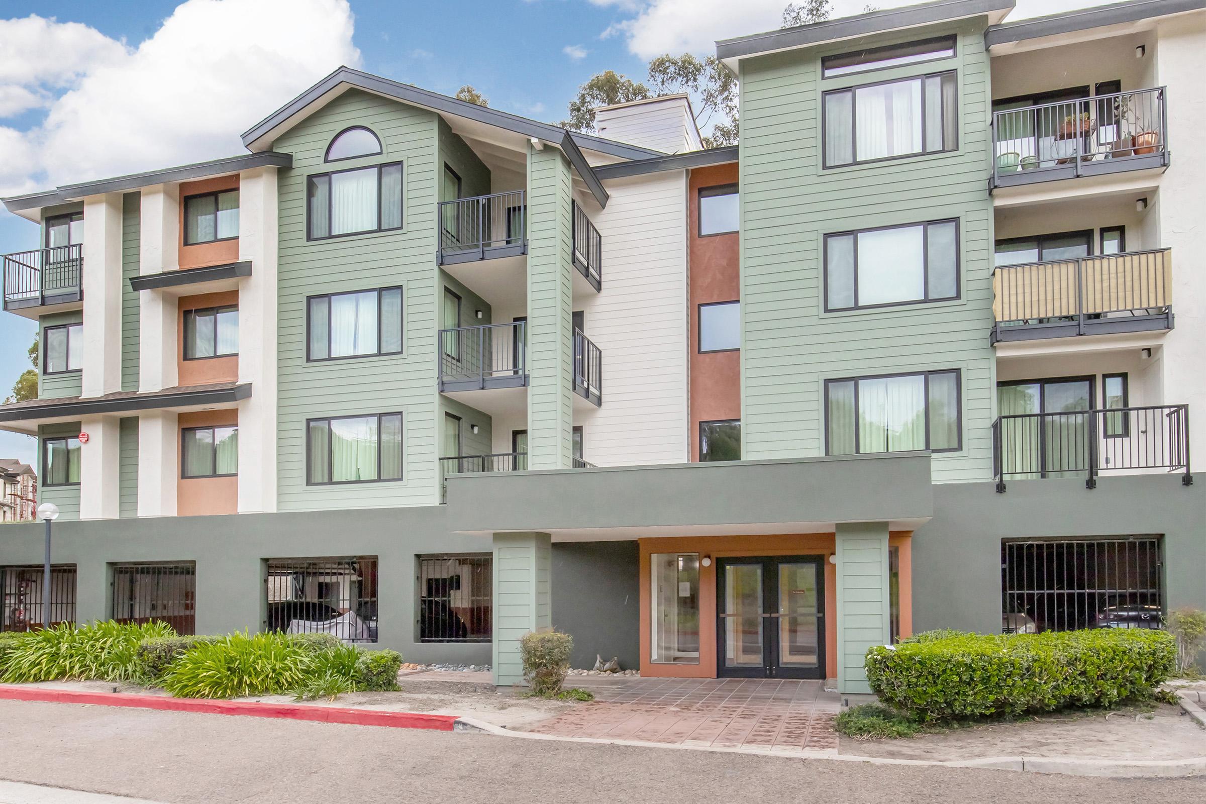 A modern multi-story building featuring a mix of green and beige exterior colors, with balconies on the upper floors. The entrance is at ground level with large windows, surrounded by landscaped greenery. The setting appears well-maintained, with a driveway leading to the entrance.