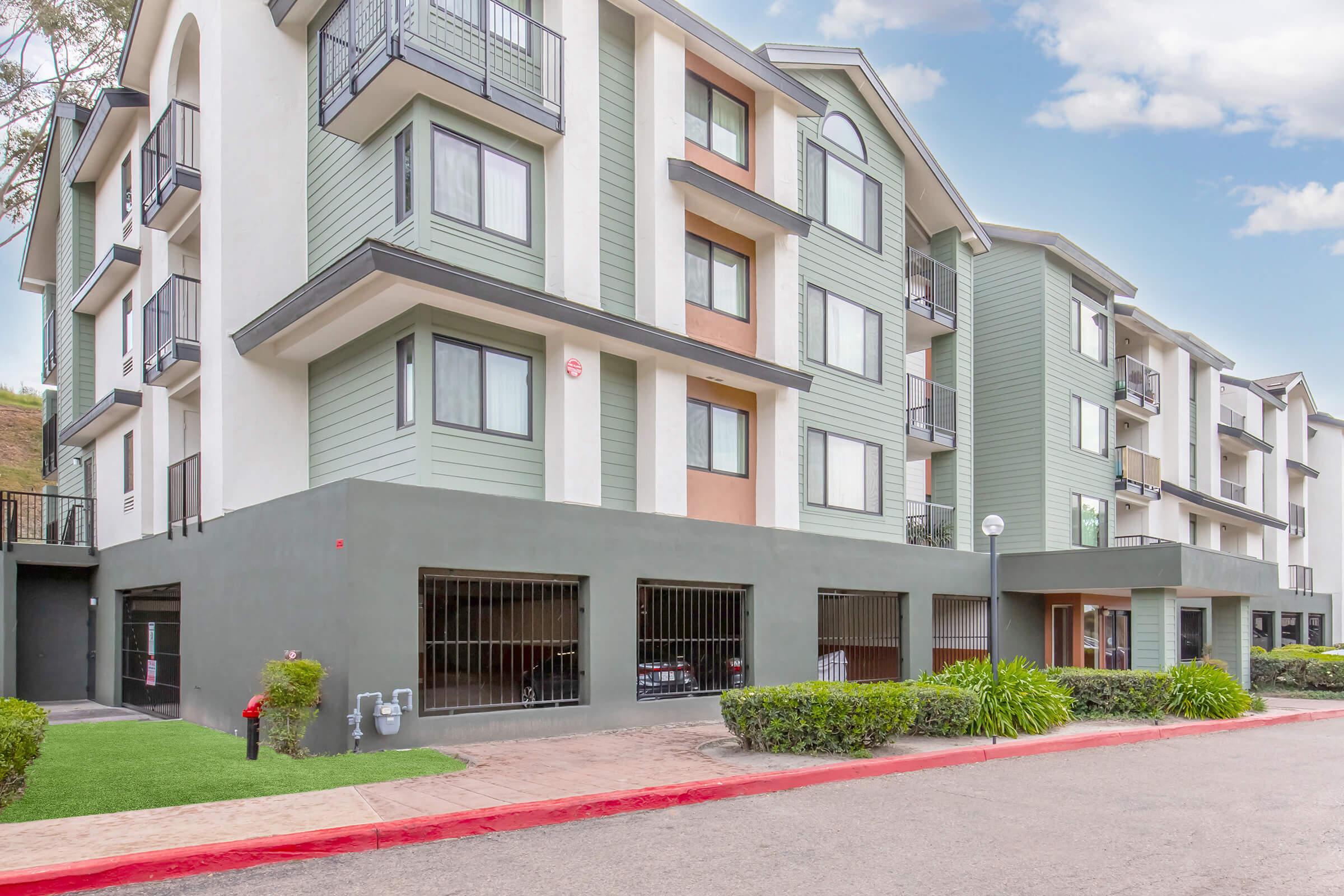 A modern multi-story apartment building featuring a combination of green and beige exterior colors. The building has balconies, large windows, and landscaped areas with grass and shrubs. A parking area with a gated entrance is visible in the foreground, along with a red curb lining the street.