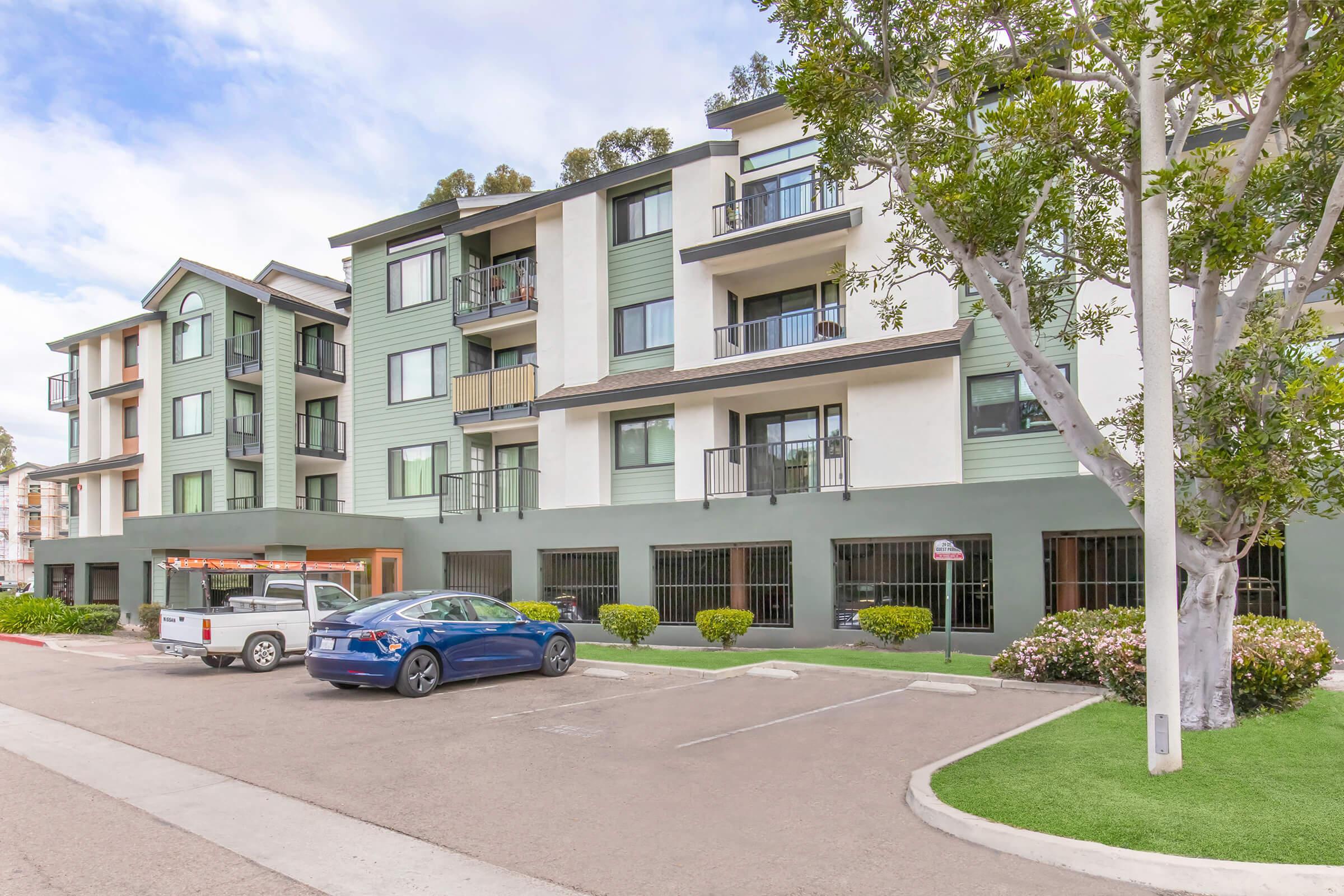 Modern apartment building with a mix of green and white exterior. The front yard features well-maintained shrubs and trees. A gray pickup truck and a blue sedan are parked in front of the building. The setting appears to be in a residential area with clear skies.