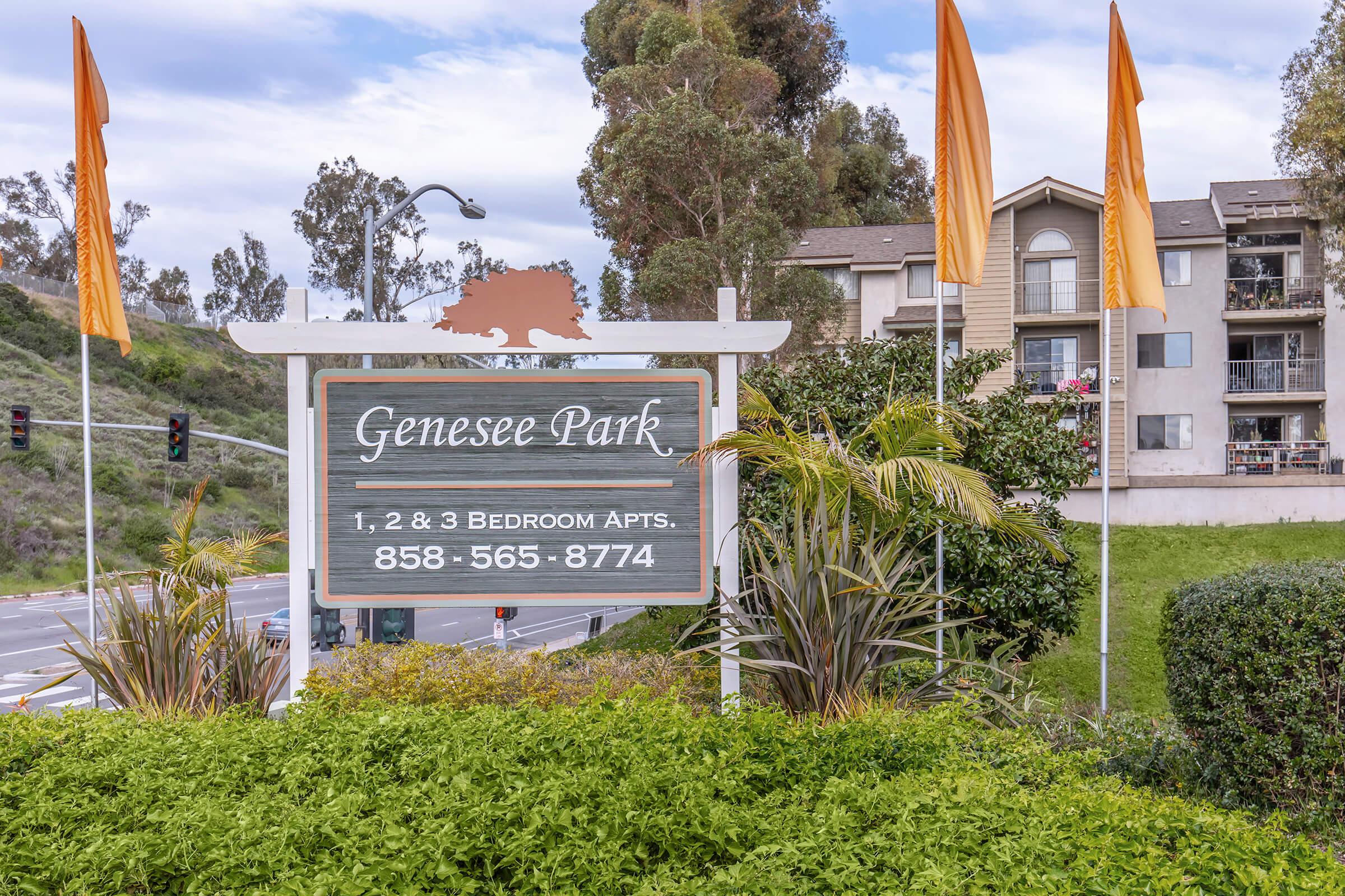 Sign for Genesee Park displaying "1, 2 & 3 Bedroom Apts" with contact number "858-565-8774". The sign is framed with plants and flags, and a building can be seen in the background, surrounded by greenery.