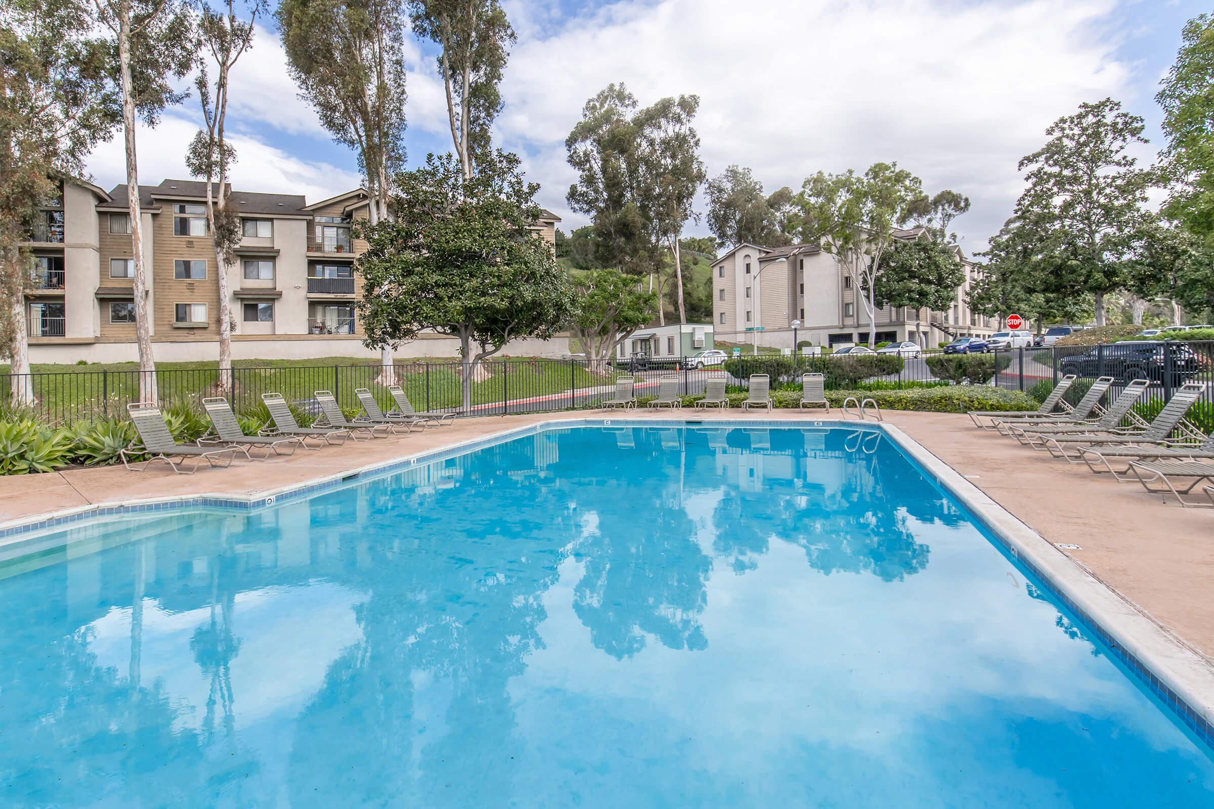 A clear blue swimming pool surrounded by lounge chairs, with tall trees and apartment buildings in the background under a partly cloudy sky. The area is well-maintained, creating a relaxing outdoor atmosphere.