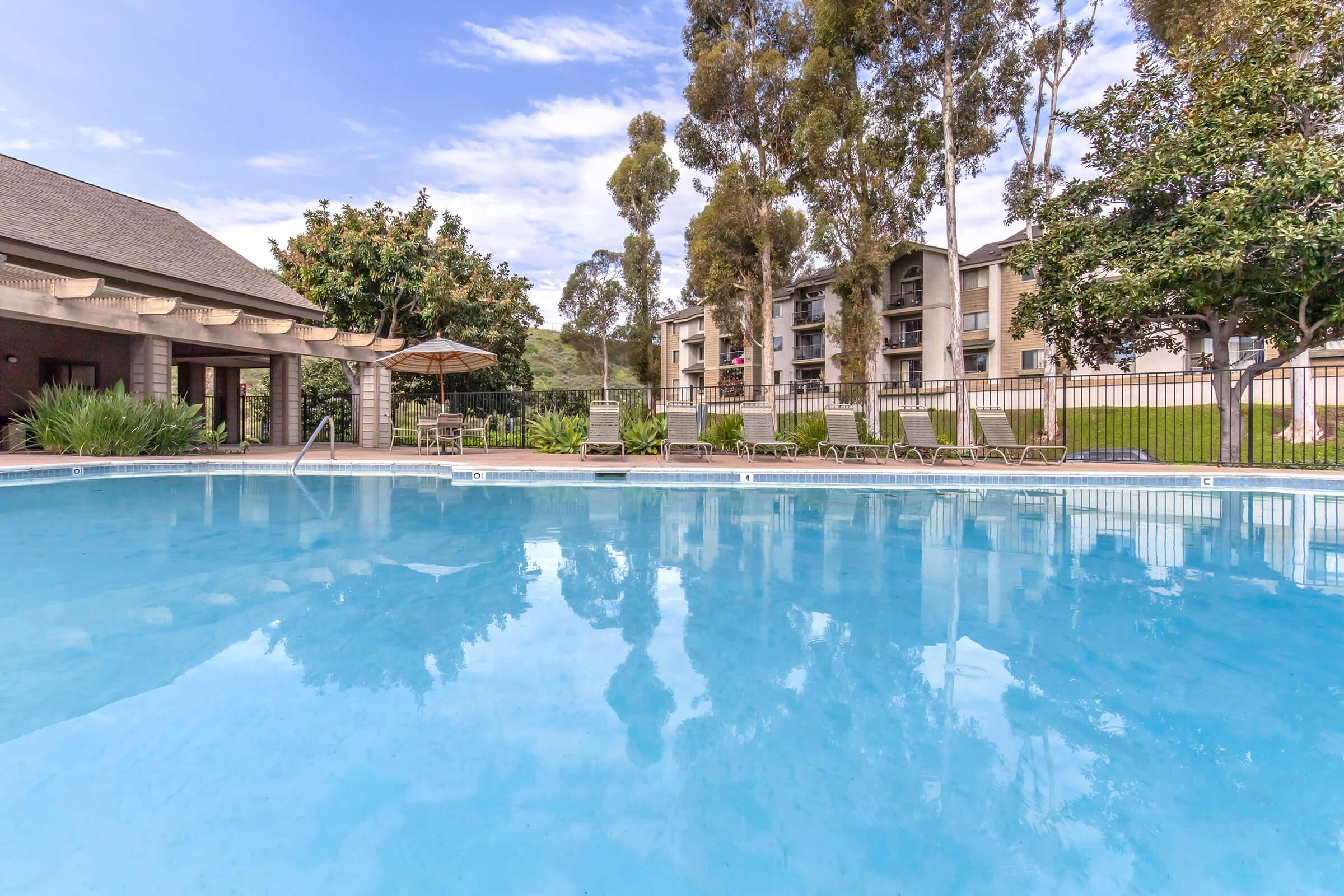 A tranquil swimming pool surrounded by lush greenery and tall trees, with a few lounge chairs nearby. Apartment buildings are visible in the background under a blue sky with scattered clouds. The water reflects the landscape, creating a serene atmosphere.