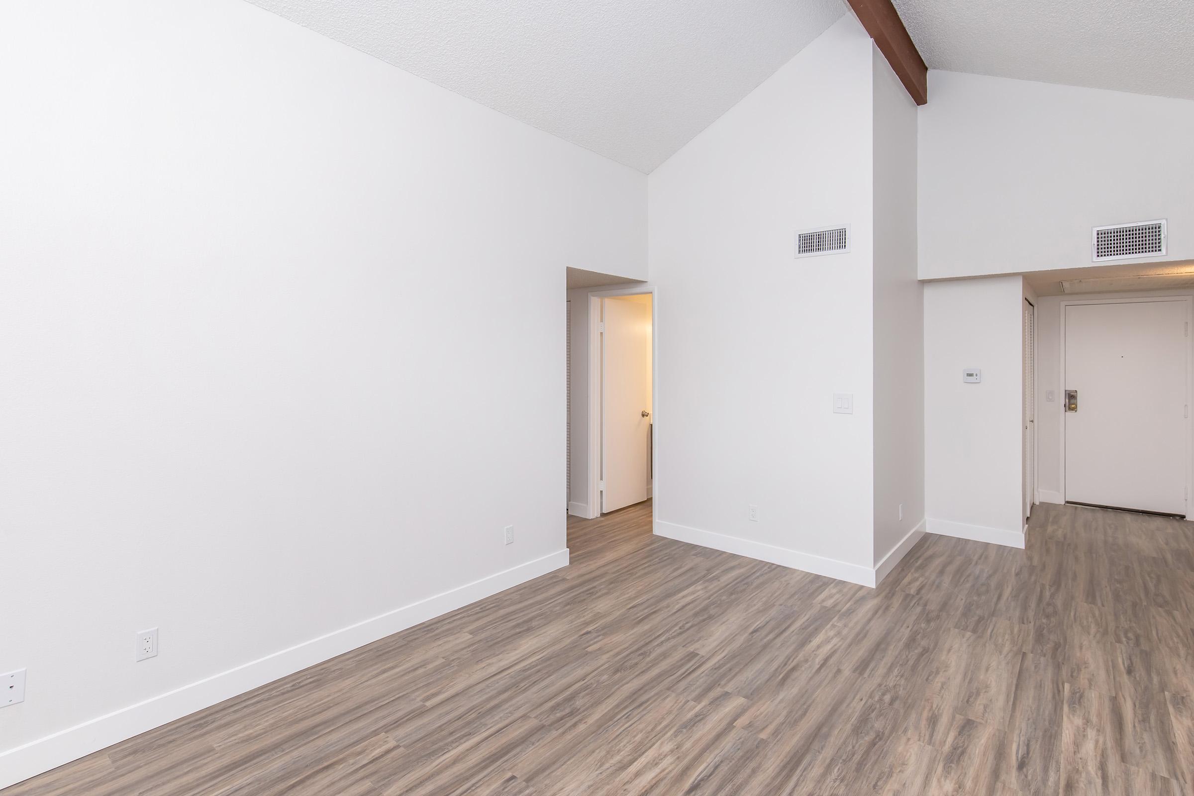 A spacious, empty room with light-colored walls and wooden flooring. The ceiling has a sloped design, and there are doorways leading to other areas. Natural light brightens the space, creating a clean and modern atmosphere.