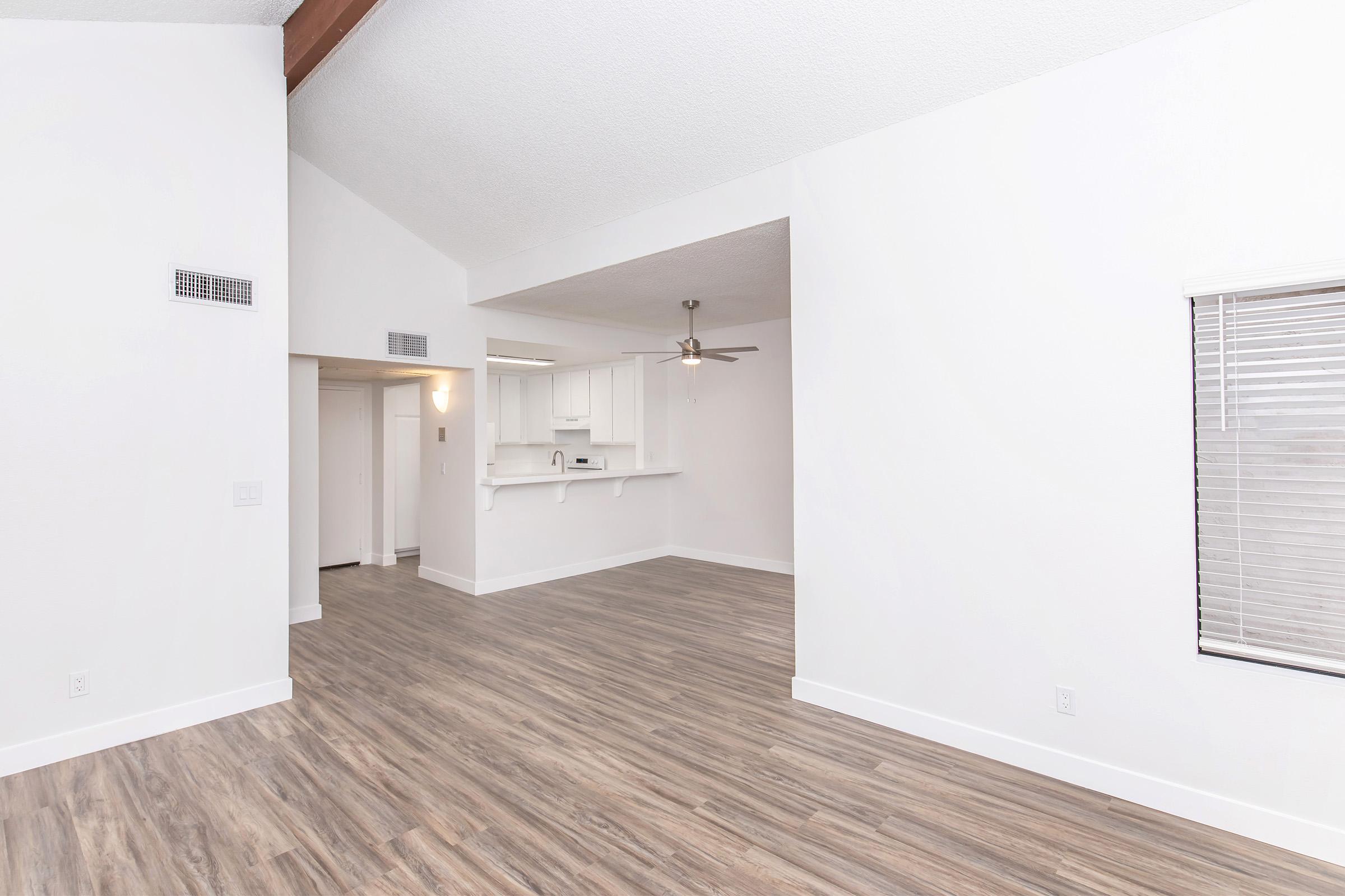 Spacious interior of a modern living room and kitchen area featuring light-colored walls, wood-like flooring, and a ceiling fan. Large windows allow for natural light, and an open floor plan connects the living space to the kitchen.