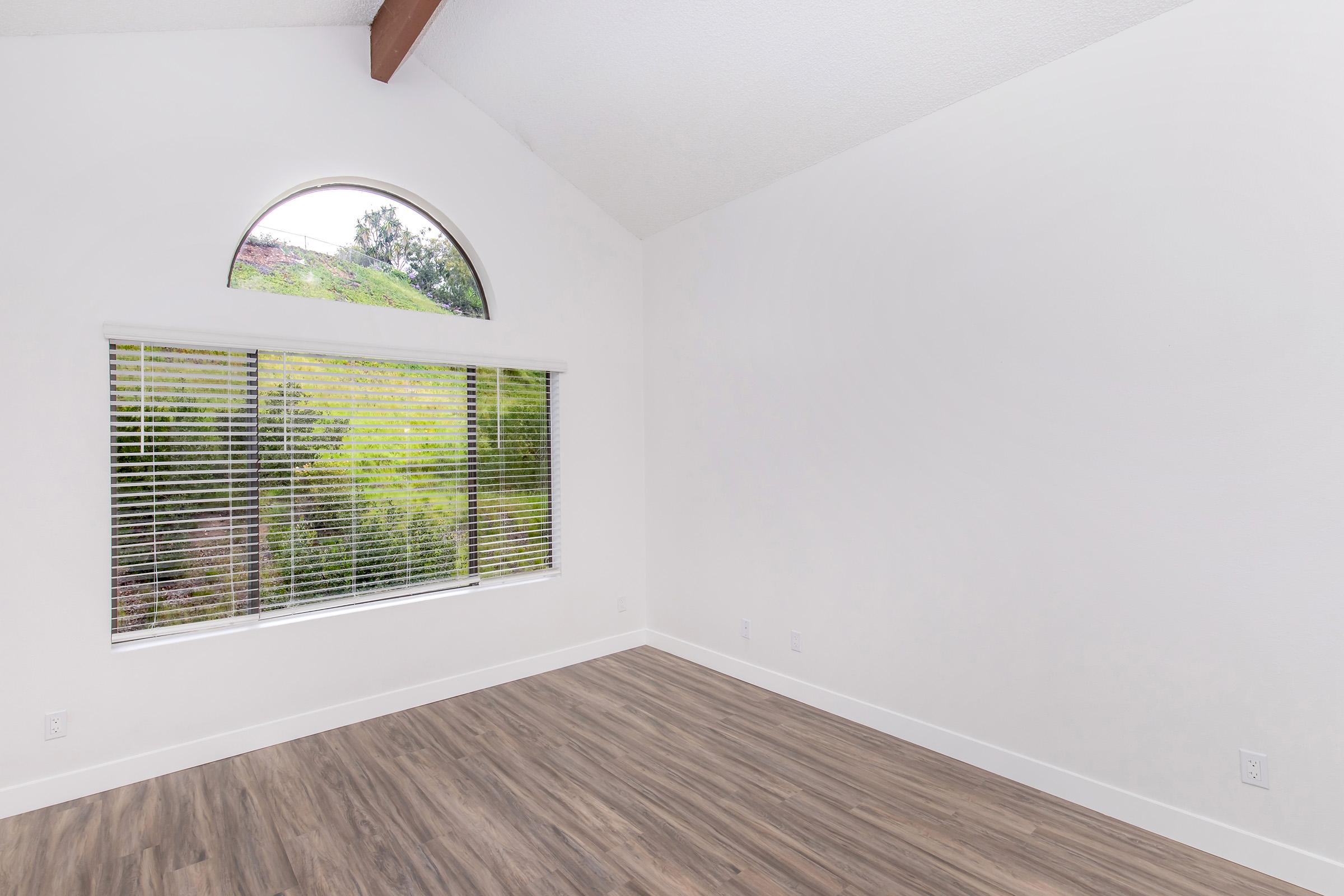 Bright, empty room with a high, arched window allowing natural light to enter. The window features horizontal blinds, and there is a view of greenery outside. The walls are painted white, and the floor is made of light wood. The room has a modern, clean aesthetic.