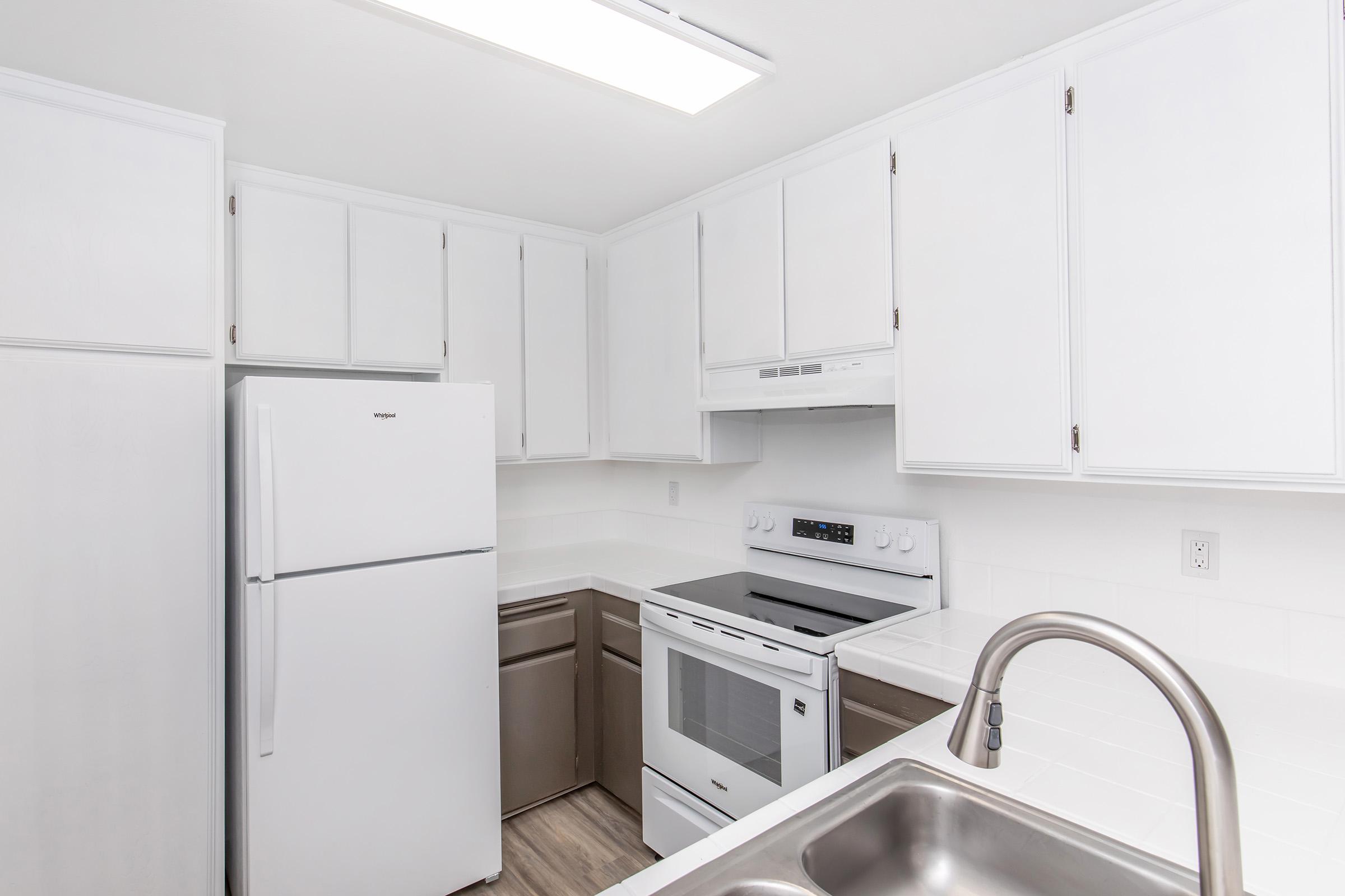 A modern kitchen with white cabinetry, featuring a stainless steel sink, a white refrigerator, an oven, and a microwave. The countertops are light-colored, and the flooring is a wood-like laminate. Natural light comes in through a ceiling fixture.