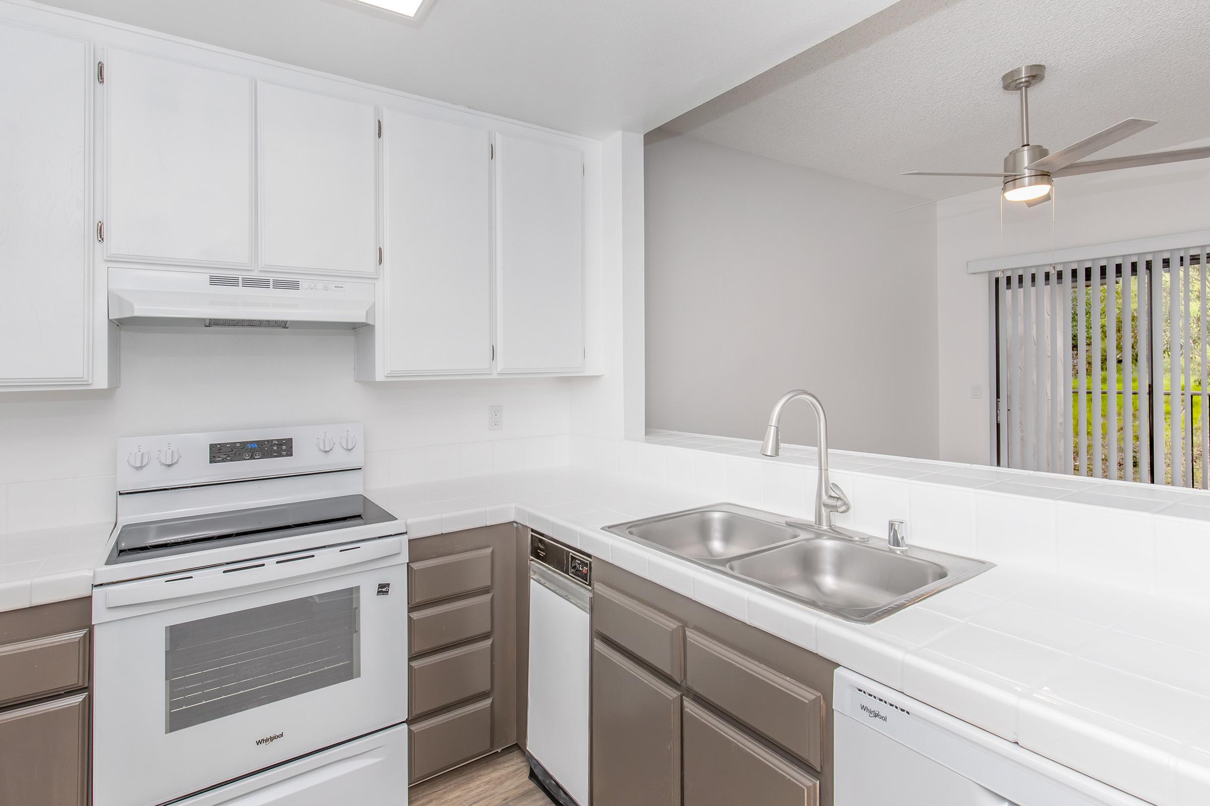 Modern kitchen featuring white cabinets and appliances, including a stove, oven, and dishwasher. A double sink is positioned between the appliances. Natural light comes through a nearby window, complemented by a ceiling fan, creating a bright and open space.