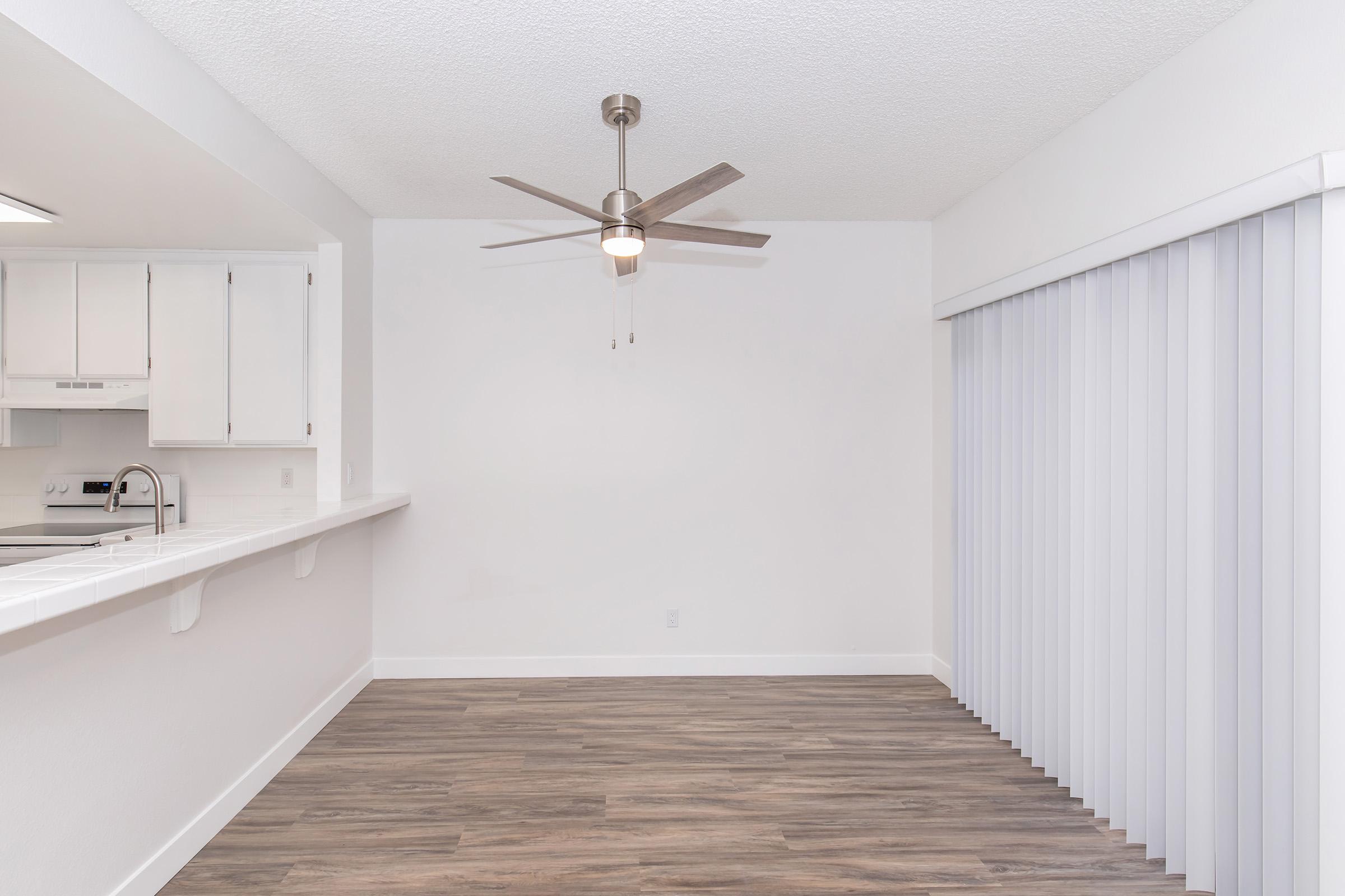A bright, modern interior featuring a ceiling fan in the center. To the left, there is a kitchen area with white cabinets and appliances. The flooring is a light wood laminate. On the right, sheer vertical blinds cover a sliding door, creating an open and airy feel in the space.