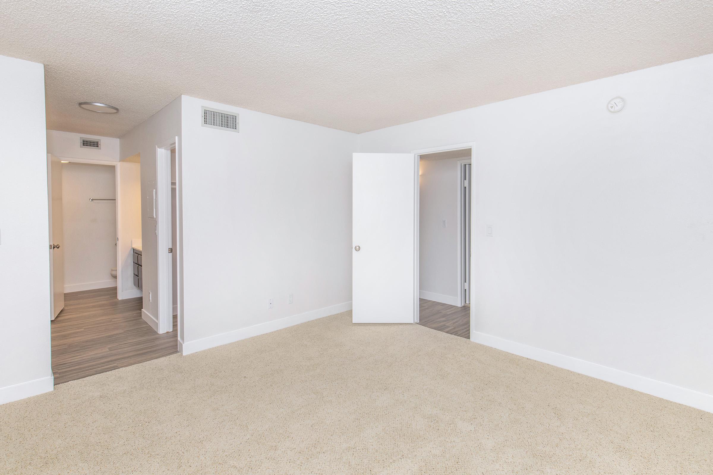 Empty room with light beige carpet, white walls, and a textured ceiling. Two doorways lead to adjacent spaces, including a bathroom. The room is well-lit, with a soft, neutral color scheme, creating a spacious and inviting atmosphere.
