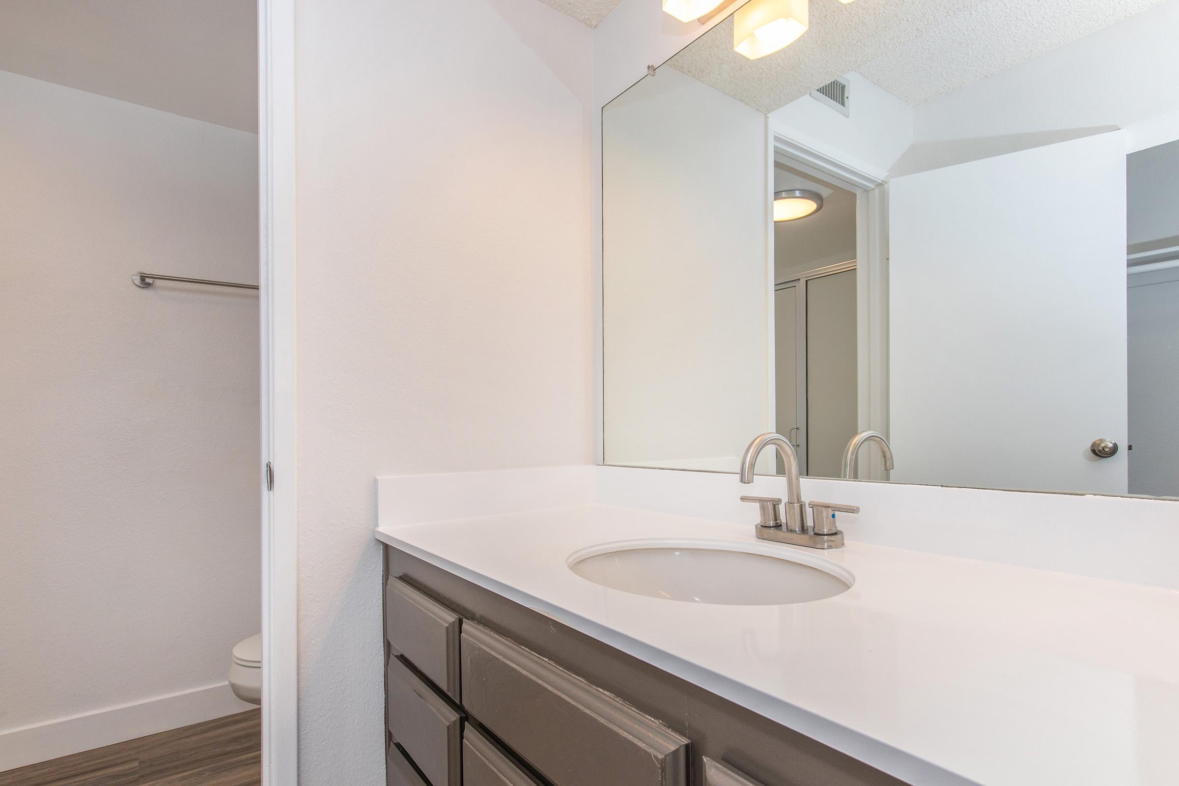 A modern bathroom featuring a white countertop with a circular sink and a sleek faucet. The walls are painted in a light color, while there is a mirror above the sink. A doorway leads to a toilet area, and another door opens to a closet space. The flooring is a light wood look.
