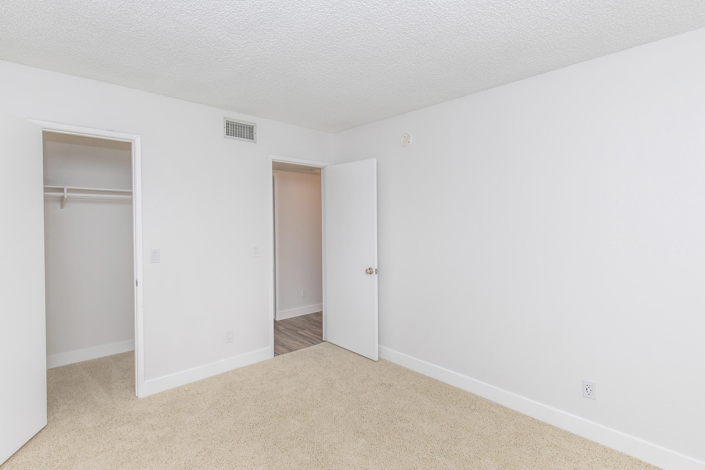 A vacant, well-lit room with beige carpet and light-colored walls. There is a closed door on one side leading to a closet, and an open door on the opposite side leading to another area. The space appears clean and unoccupied, suitable for various purposes.