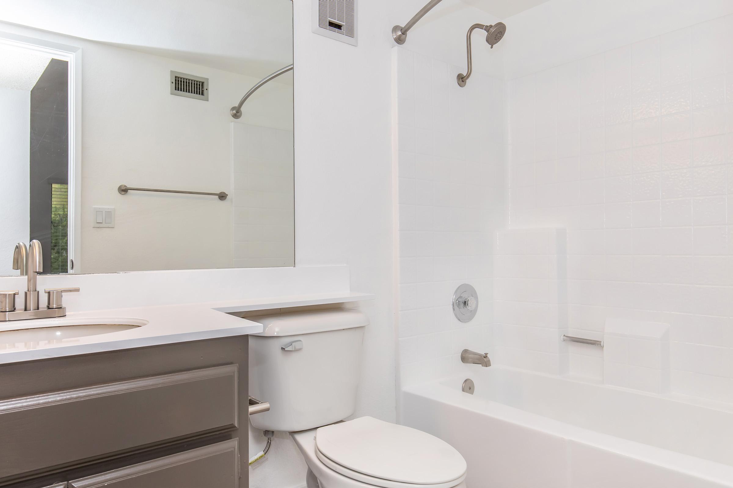 A clean, modern bathroom featuring a white bathtub with a shower fixture, a large mirror above a simple sink with gray cabinets, and a white toilet. Bright lighting enhances the fresh, minimalist design, with a doorway visible in the background leading to another room.