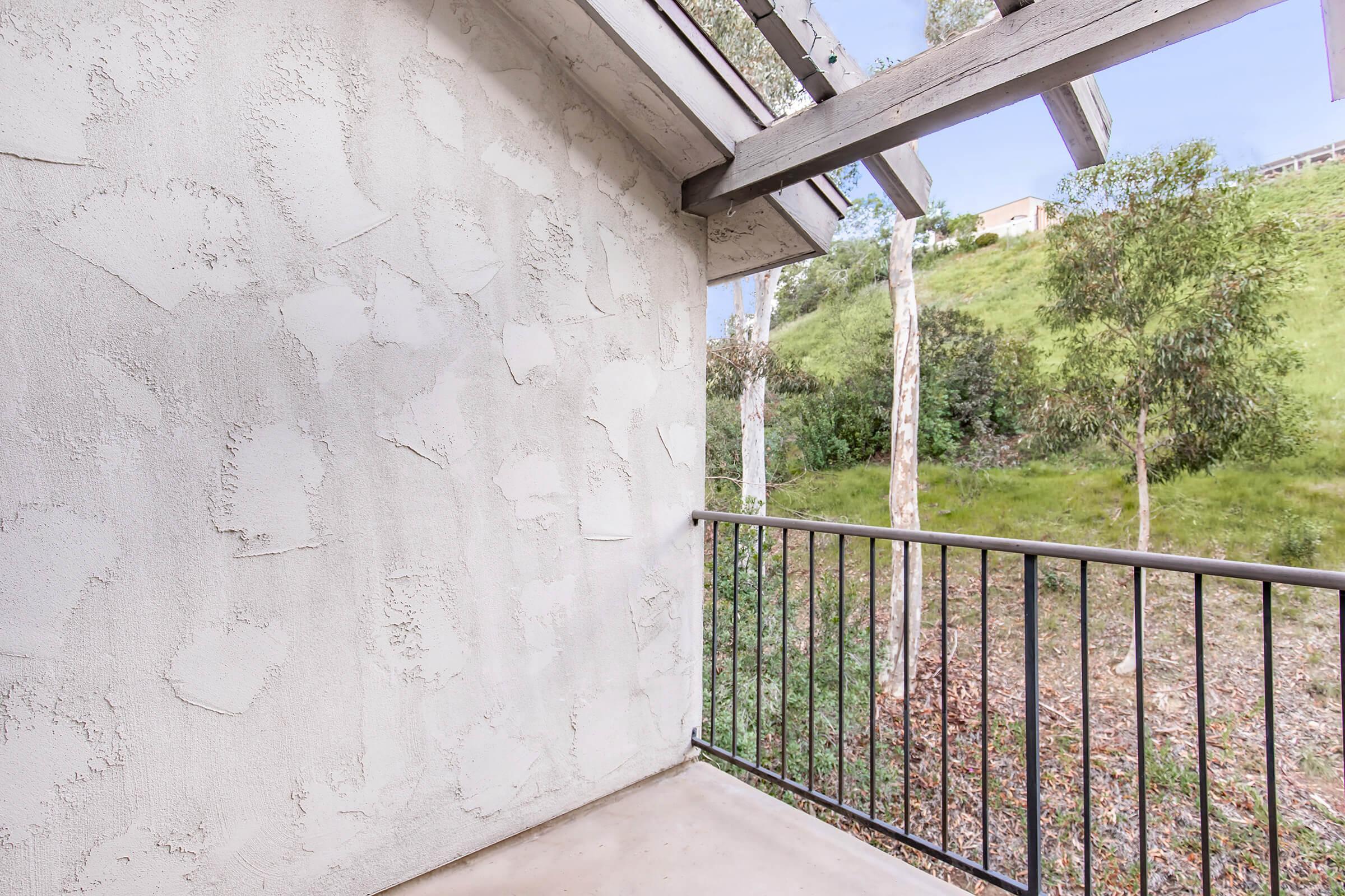 A small balcony with a black metal railing overlooks a green hillside filled with trees. The wall of the balcony is textured, and the setting appears peaceful with natural surroundings.
