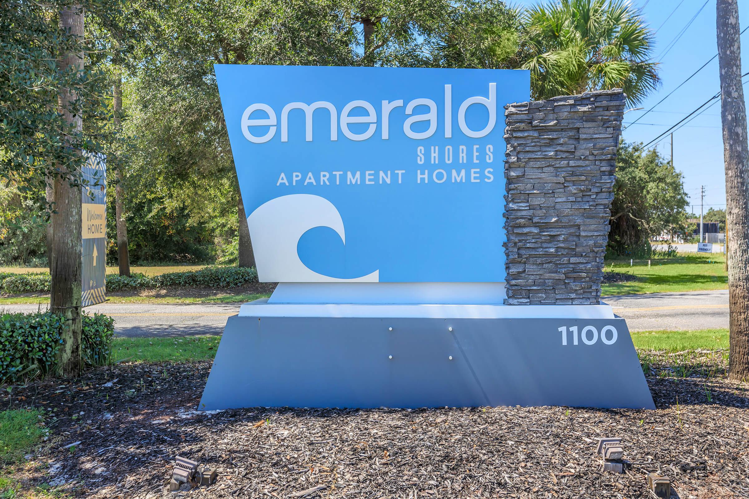 Sign for Emerald Shores Apartment Homes, with a stylized wave graphic and stone design. The location is marked with the number "1100." The background features greenery and trees, indicating a residential area.