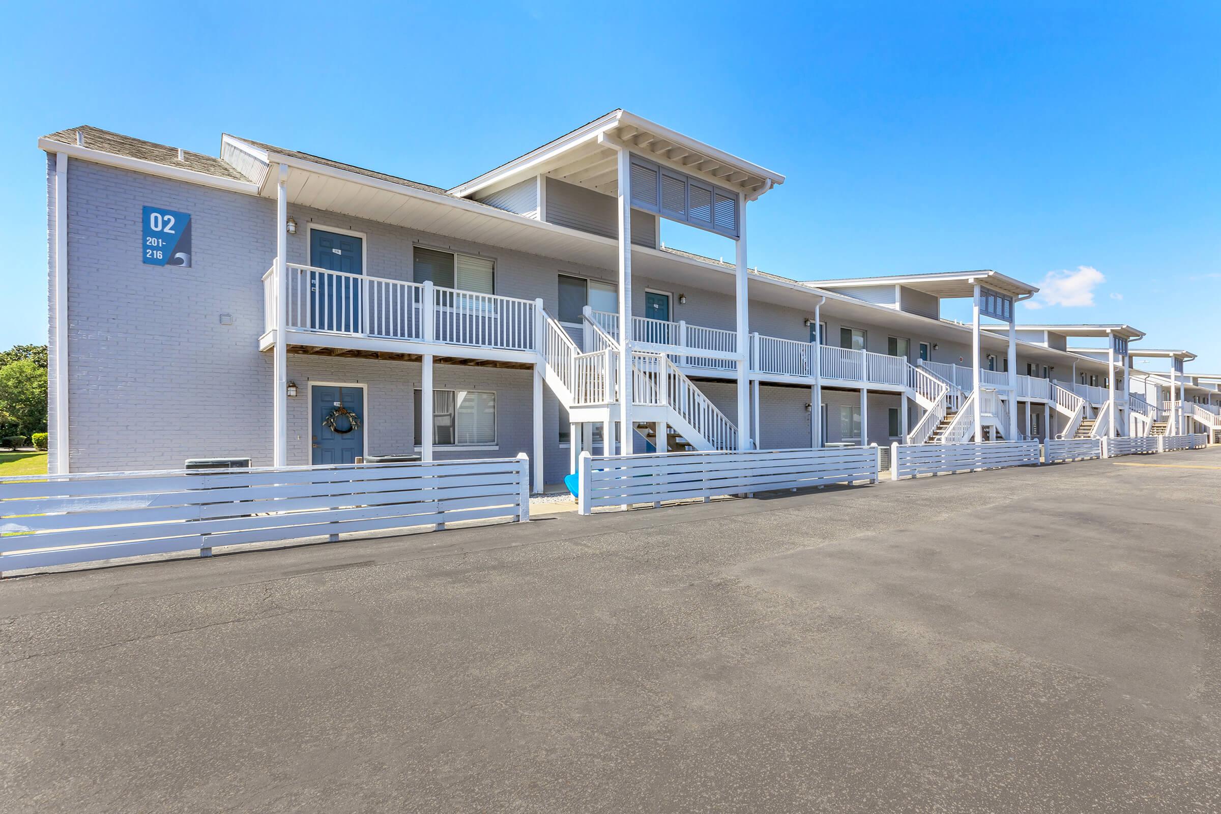 A view of a two-story building with a modern design featuring multiple units. Each unit has a balcony and a white railing, with stairs leading to the upper level. The parking area is in the foreground, and the sky is clear with a few clouds. The building has a light color scheme, contributing to a fresh appearance.