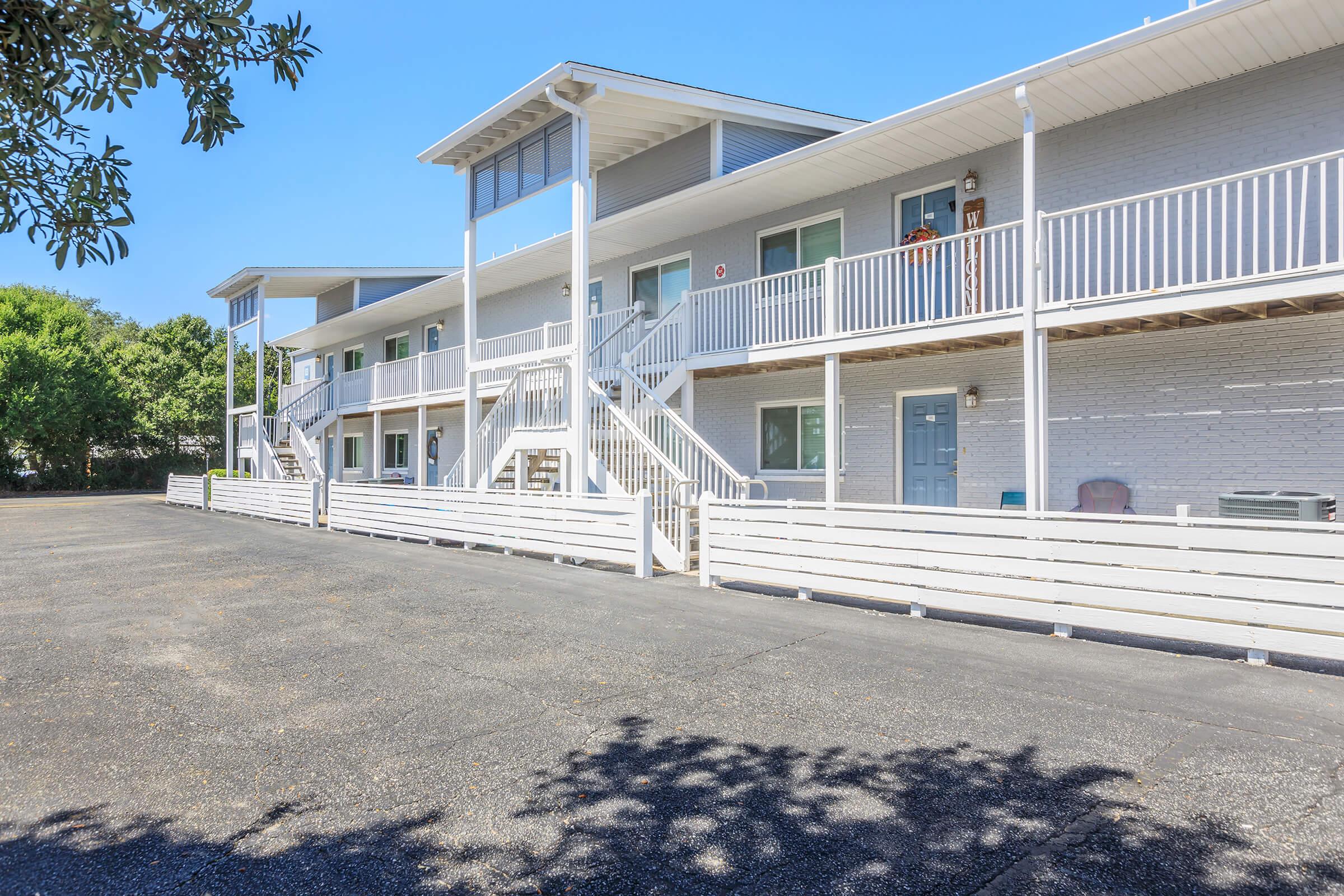 A view of a multi-story motel with a white exterior. The building features balconies, stairs, and several entrances. In the foreground, there is a paved parking area surrounded by greenery and trees in the background under a clear blue sky.