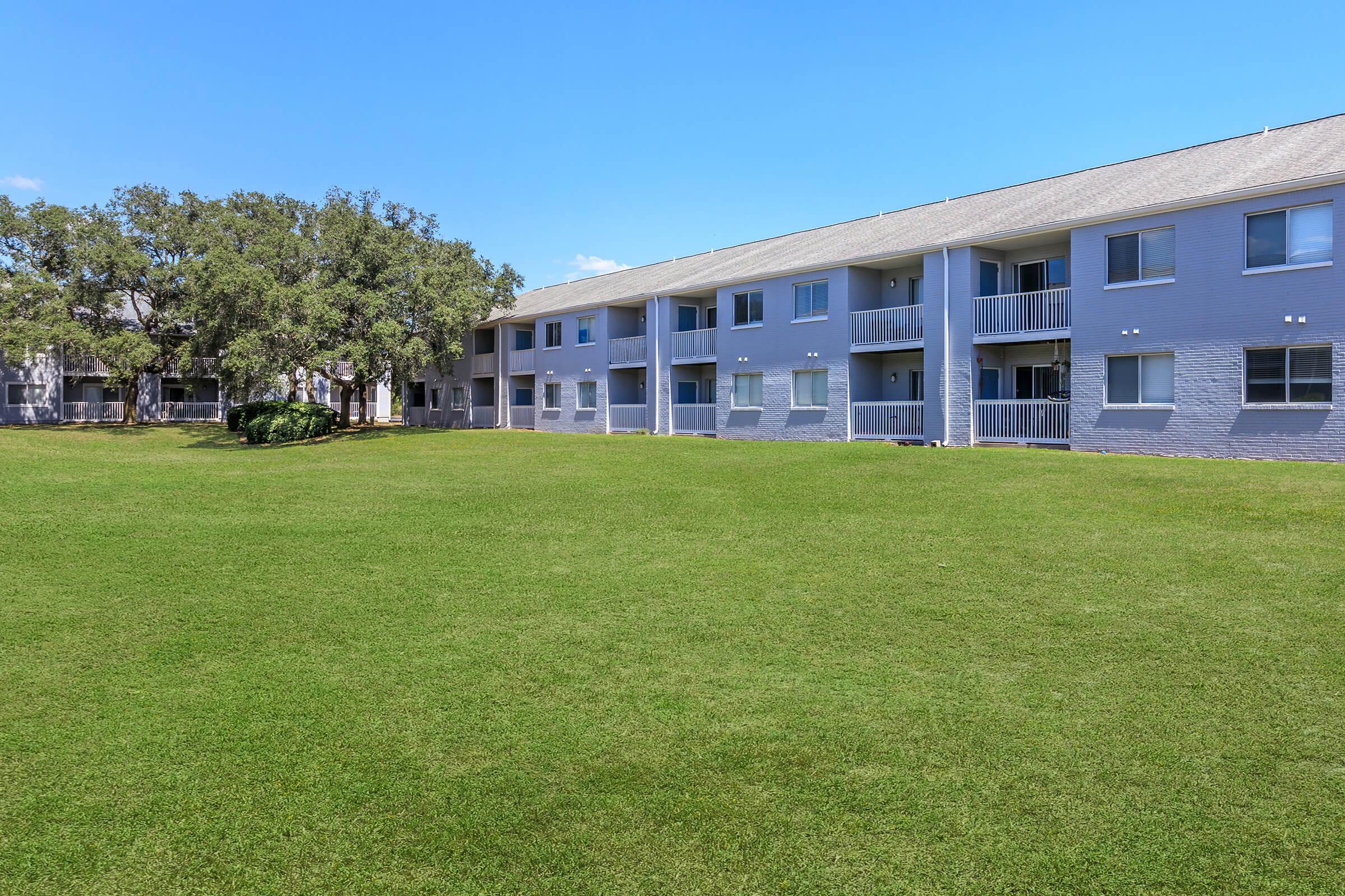 A grassy area in front of a light-colored, two-story apartment building with balconies. The building features several windows and is surrounded by trees under a clear blue sky. The scene conveys a peaceful residential atmosphere.