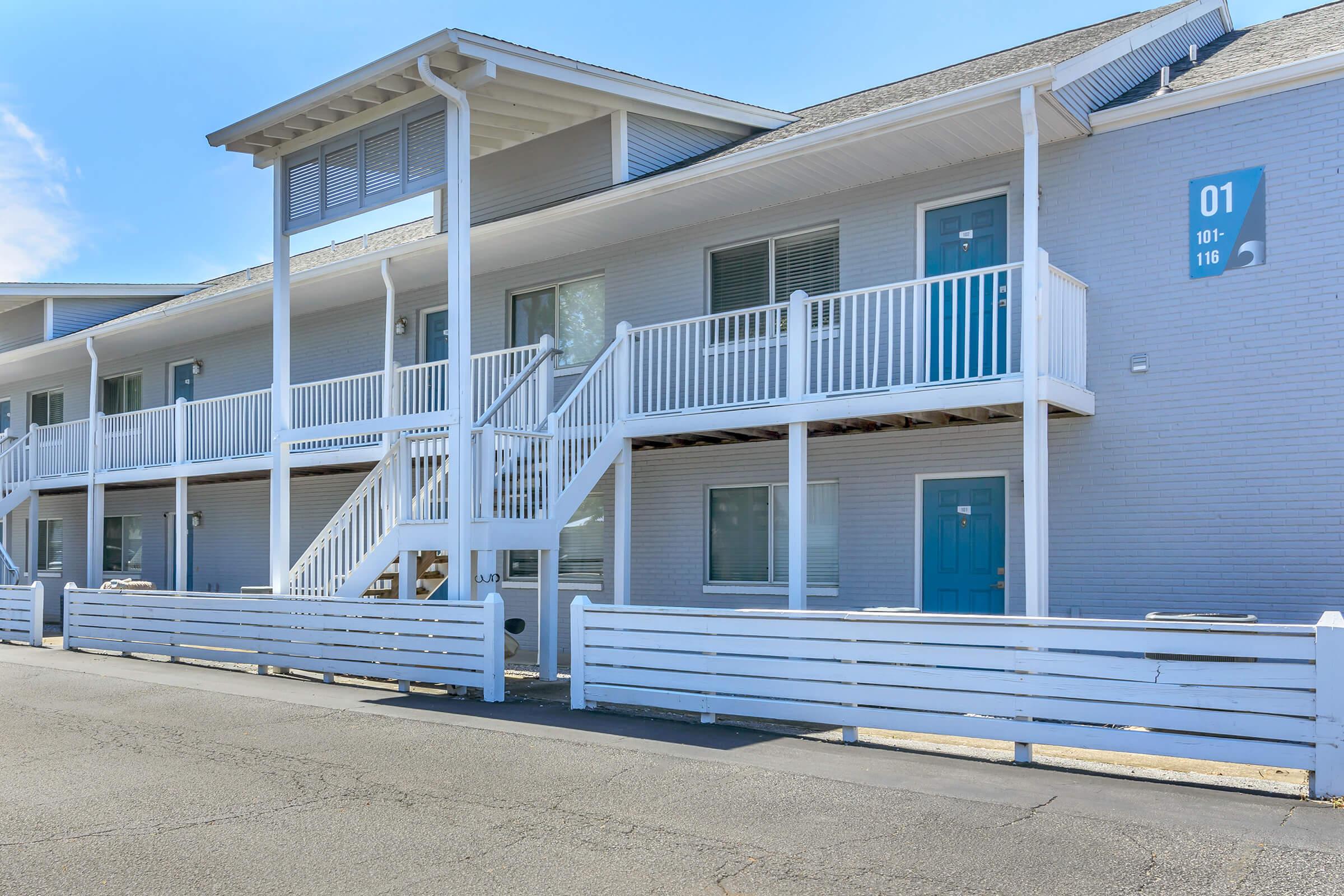 A white multi-unit building with a staircase leading to the second floor. The building features blue doors and large windows, with a white fence in front. The sky is clear and blue, indicating a sunny day.