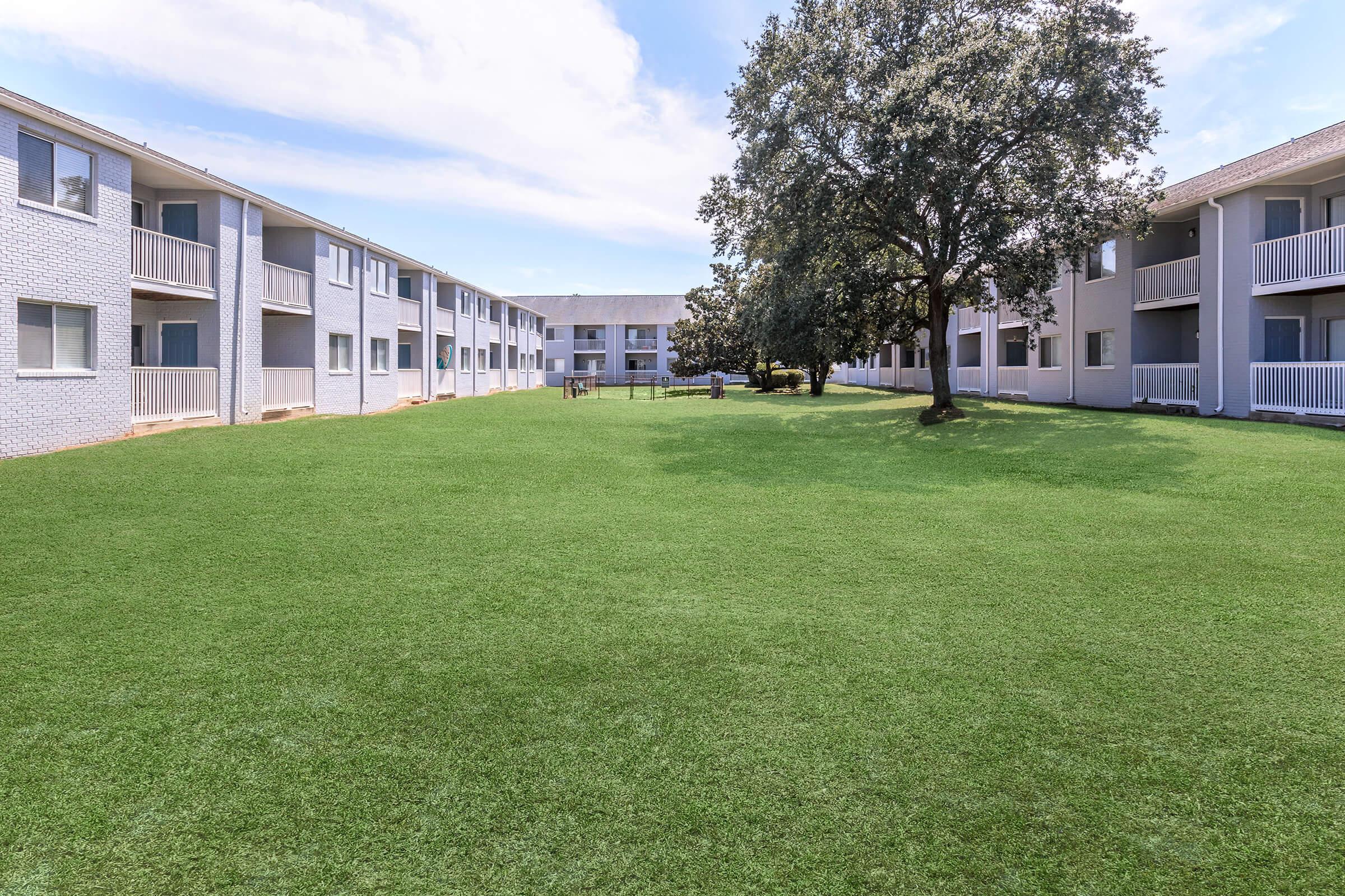 View of a grassy courtyard surrounded by two sets of apartment buildings. The area is well-maintained with a large, bright lawn and a few trees in the background, under a clear blue sky with some clouds. The architecture features multiple balconies, creating a relaxed residential atmosphere.