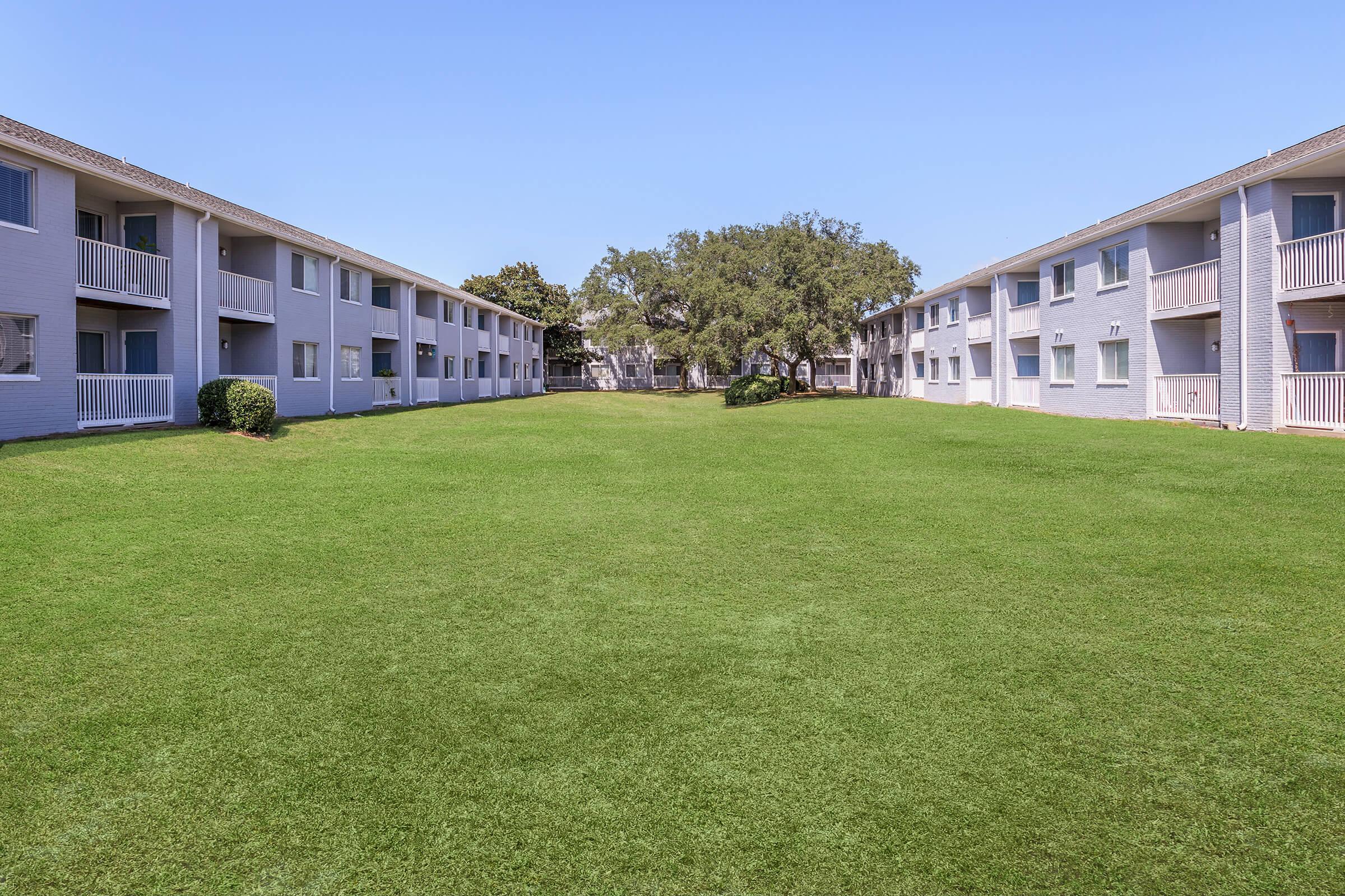 A landscaped courtyard view between two apartment buildings, featuring well-maintained lawns and greenery. The buildings are light-colored with balconies, surrounded by trees, creating a peaceful environment. The sky is clear and blue, enhancing the serene atmosphere.