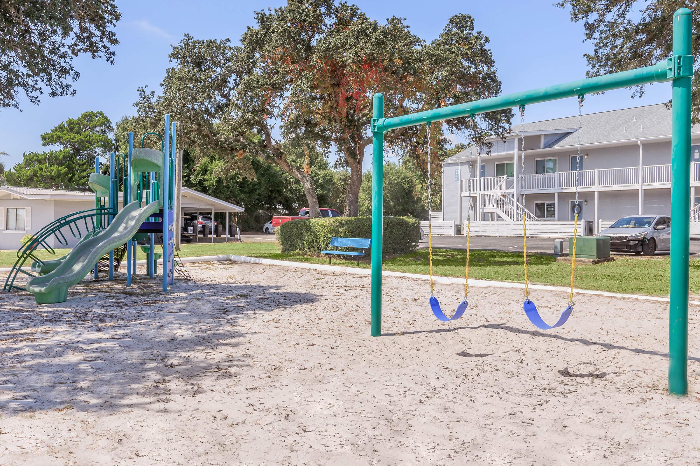 A bright playground scene featuring a green slide and climbing structure, surrounded by sandy ground. Two swings hang from a frame, and in the background, there are residential buildings and a parked car. Lush trees provide shade, creating a cheerful outdoor space for children to play.