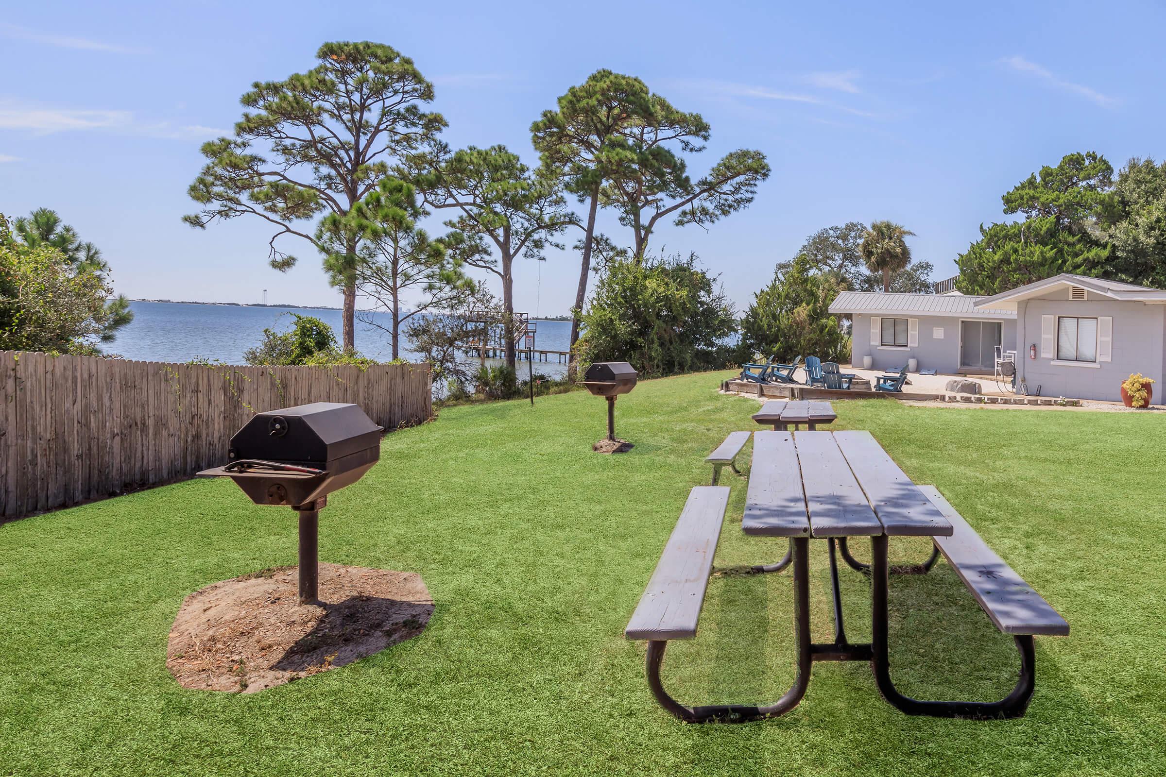 A scenic outdoor space featuring a picnic area with wooden tables, charcoal grills, and a view of a calm water body. Lush green grass and trees line the area, with a house visible in the background, creating a relaxing atmosphere for gatherings and barbecues.