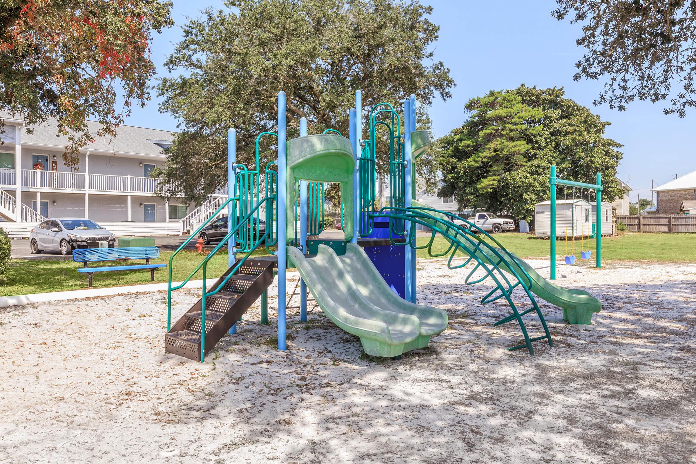 A colorful playground featuring green slides, climbing structures, and a staircase, surrounded by grassy areas and trees. Nearby, a blue bench is visible on a sandy surface, with buildings and vehicles in the background. The setting is bright and inviting for children to play.