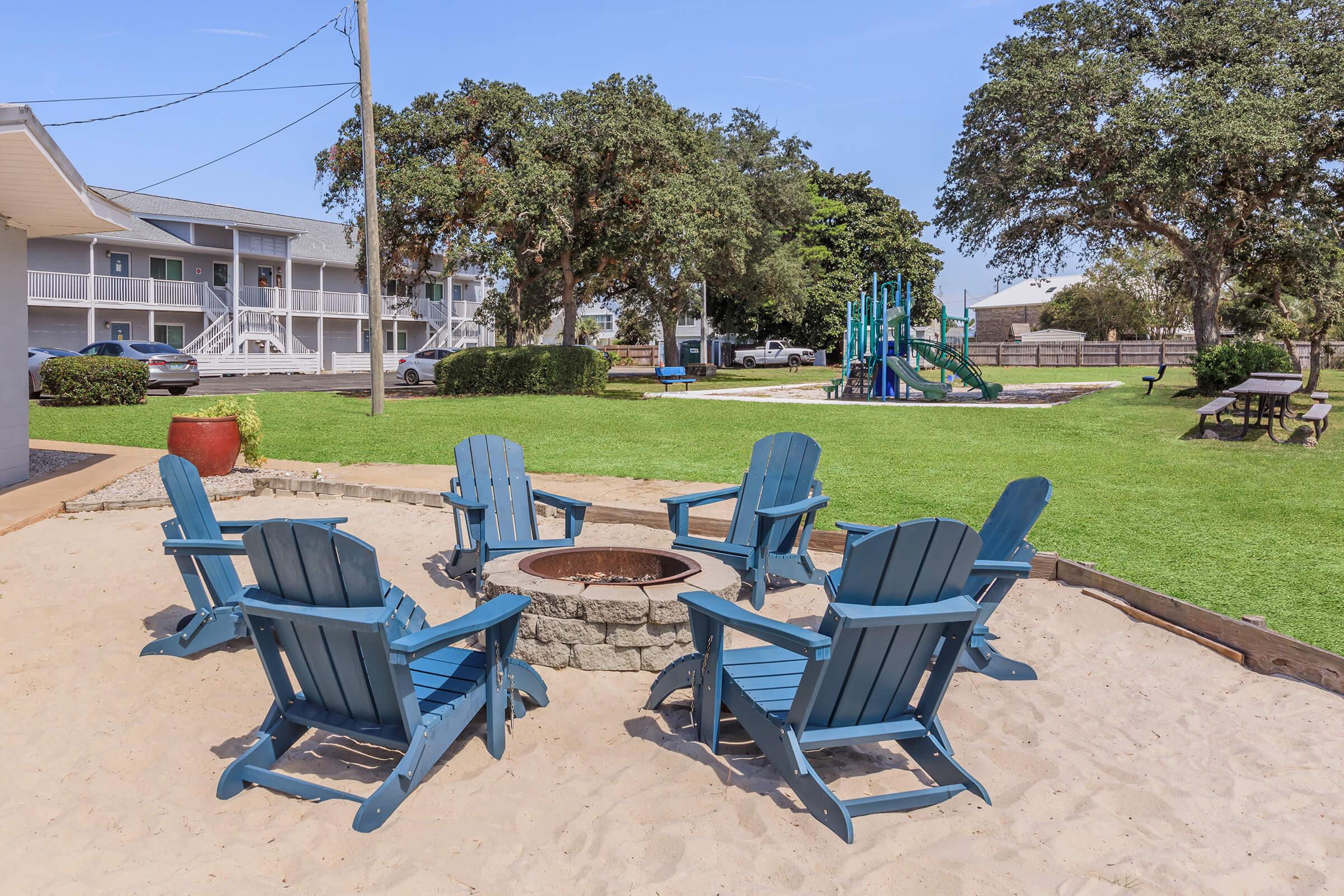 A sandy fire pit area with blue Adirondack chairs arranged in a circle, surrounded by a grassy lawn. In the background, there are multi-story buildings and a playground with slides and climbing equipment. Large trees provide shade in the outdoor setting.