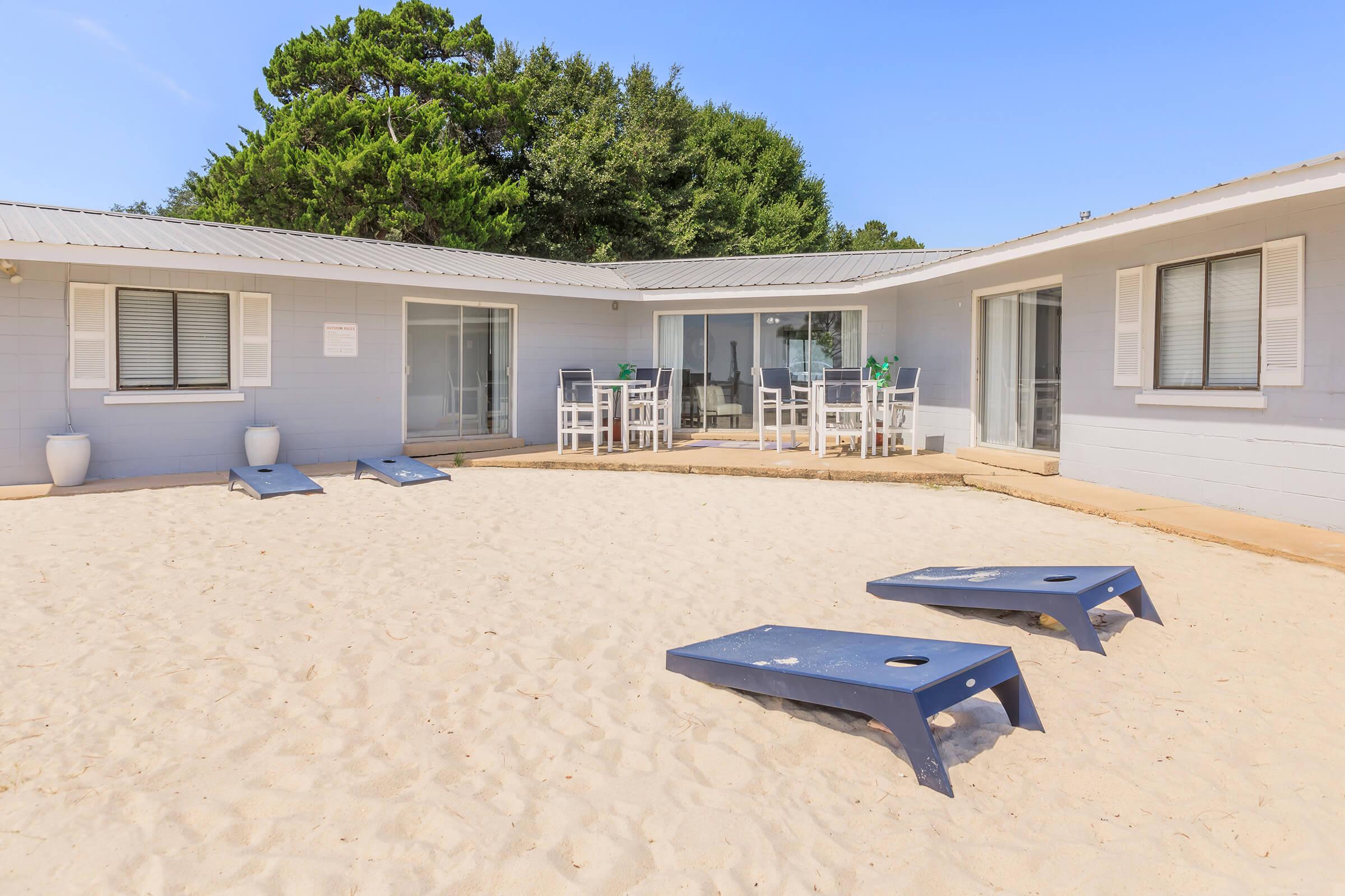 A sunny beach house patio with white sandy ground. Two blue lounge chairs are positioned in the sand. In the background, there's a light gray house with large windows and a dining area, surrounded by trees under a clear blue sky.