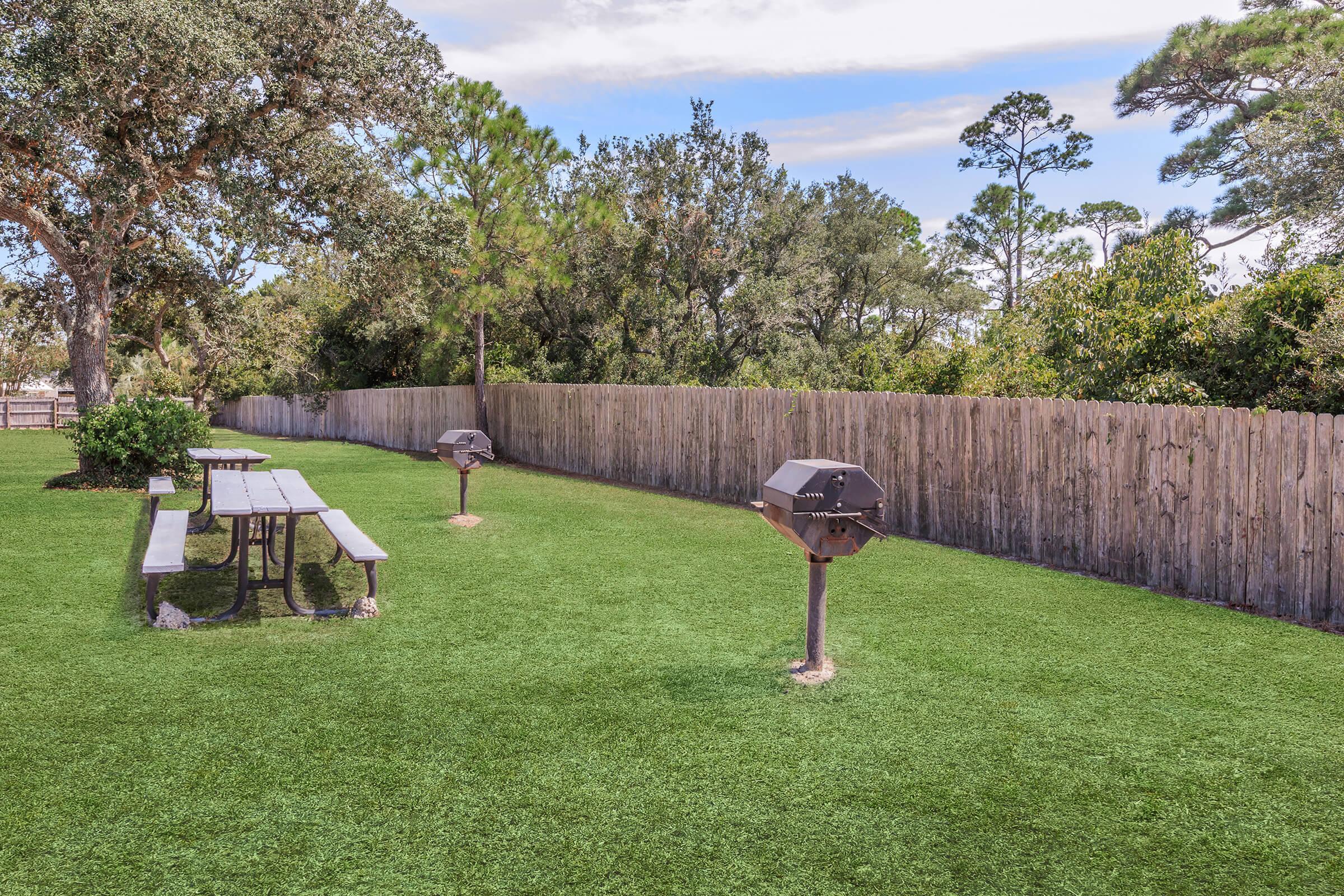 A grassy outdoor area with a wooden picnic table and two charcoal grills. The space is enclosed by a wooden fence and surrounded by trees, creating a peaceful park-like setting. The sky is partly cloudy, adding to the serene atmosphere.