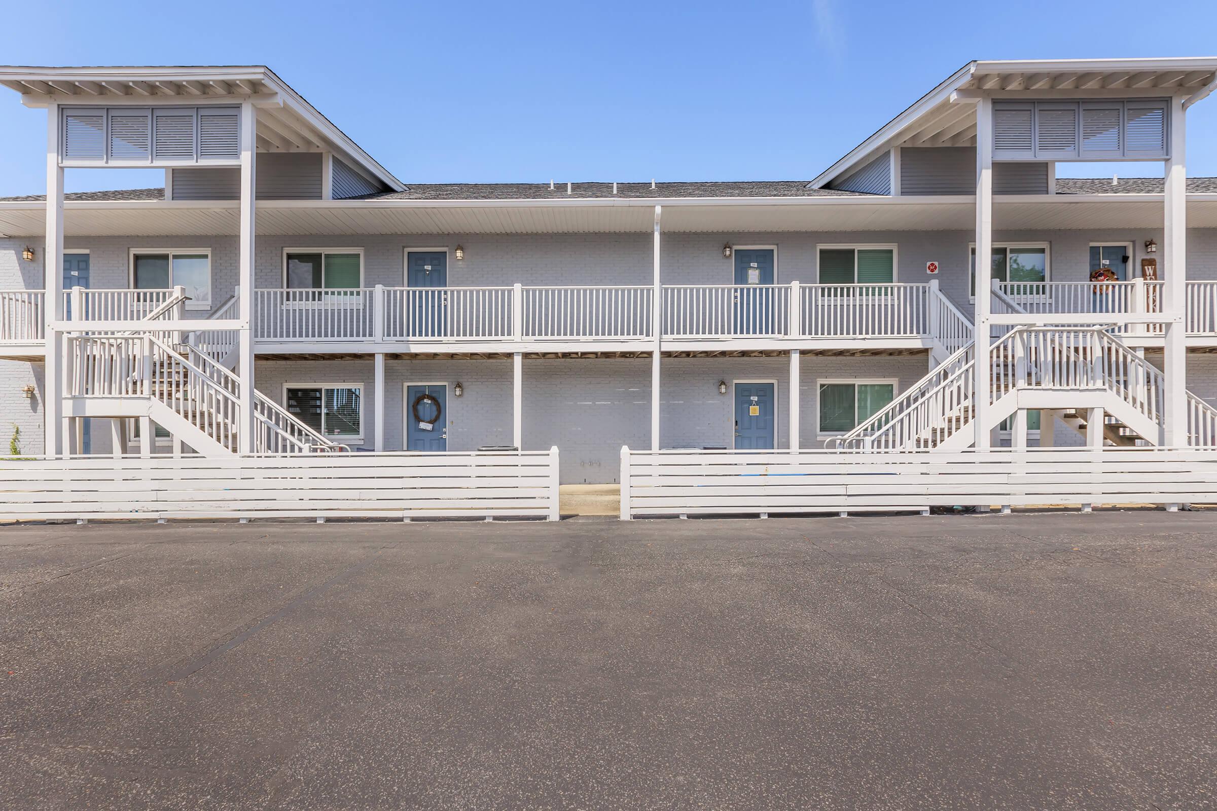 A two-story building with multiple hotel-style rooms, featuring white railings and a light-colored exterior. Each room has a door facing a central walkway with stairs on either side. The parking area is visible in front, with clear blue skies in the background.