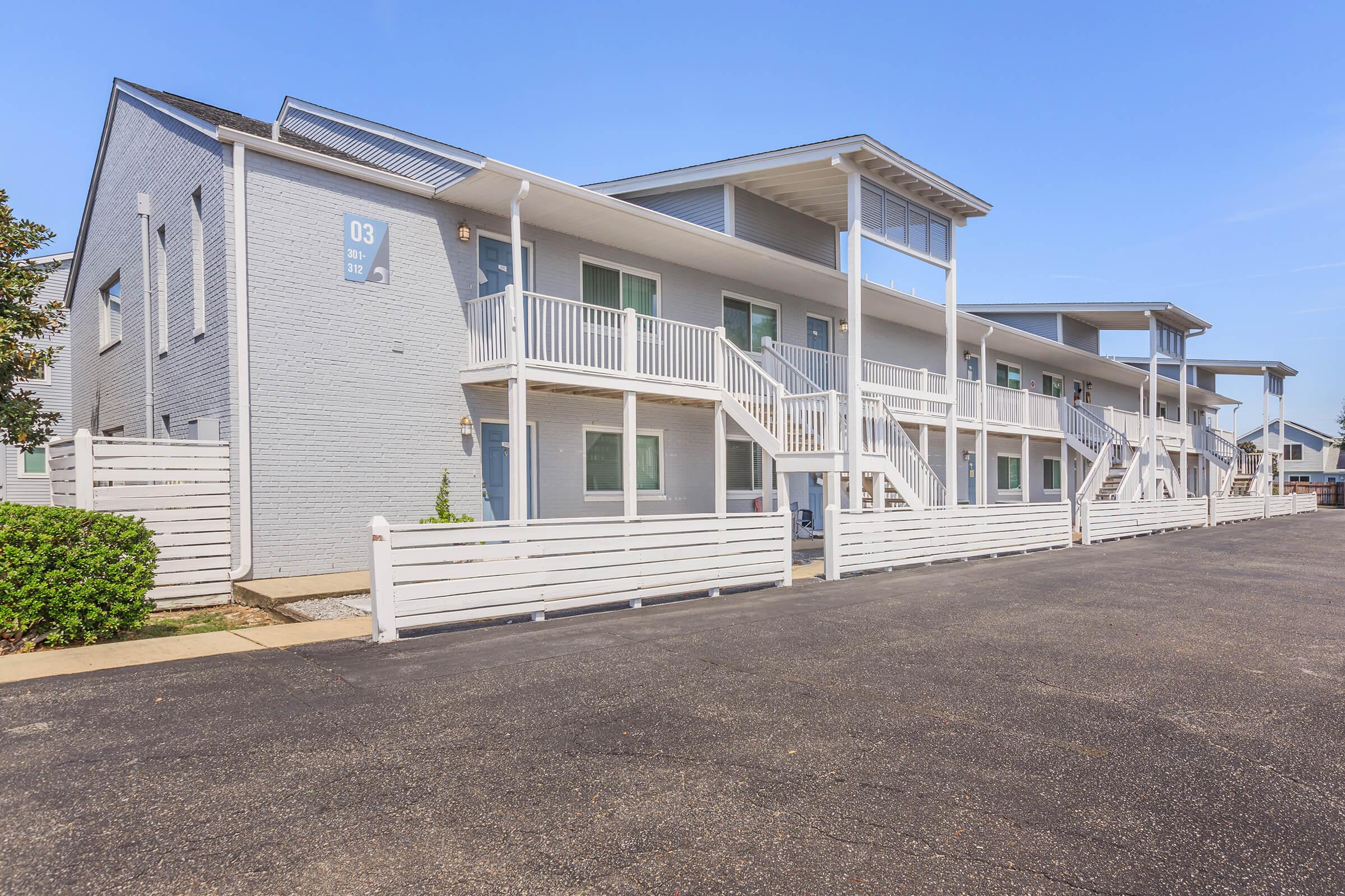 A view of a modern, two-story building with multiple balconies and white railings. The exterior features light gray siding and a clear sky above. There's a paved parking area in front, along with a low white fence and some greenery. The setting suggests a residential or vacation property.