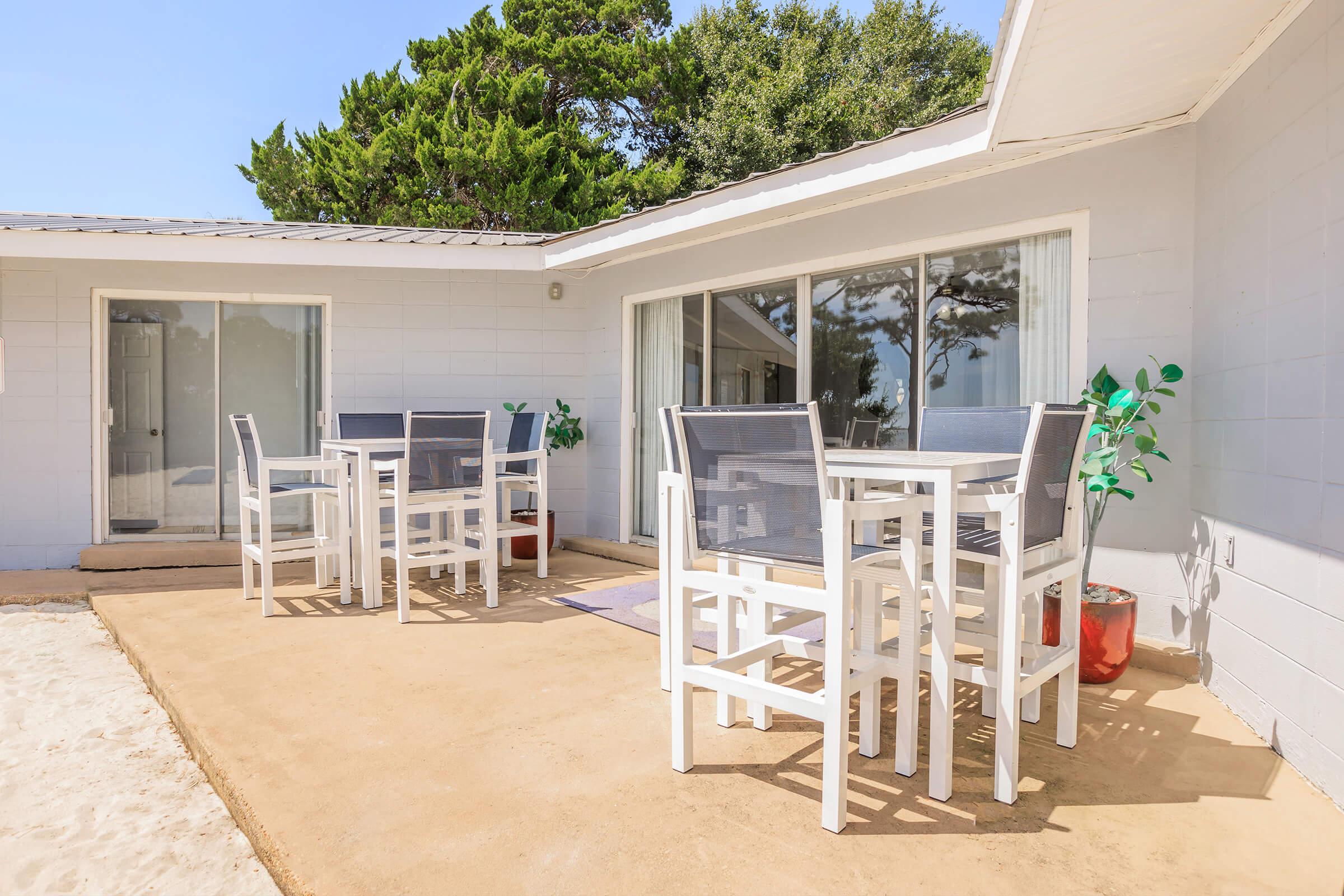Outdoor patio area featuring several white high tables and chairs, surrounded by sand and greenery. Large sliding glass doors provide access to an indoor space, with a vibrant potted plant adding a touch of color. The setting is bright and inviting, ideal for relaxation or dining.