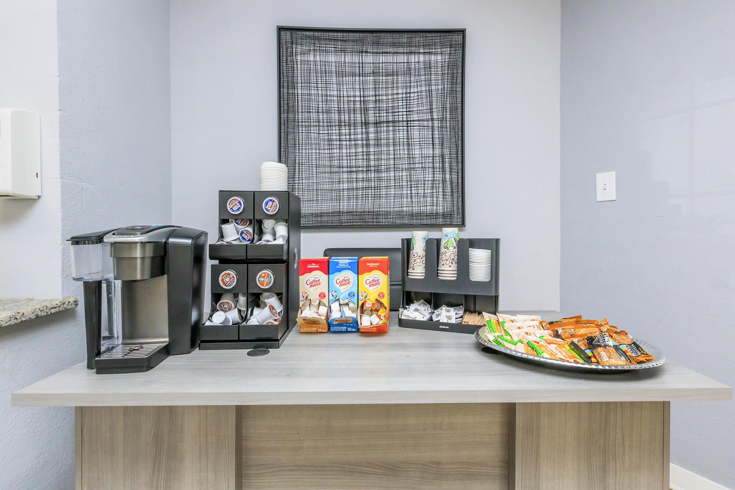 Coffee station with a Keurig machine, various flavored coffee pods, and creamers displayed alongside cups. A tray of assorted snacks, including granola bars and pastries, is set on a gray countertop, with a patterned wall hanging in the background.
