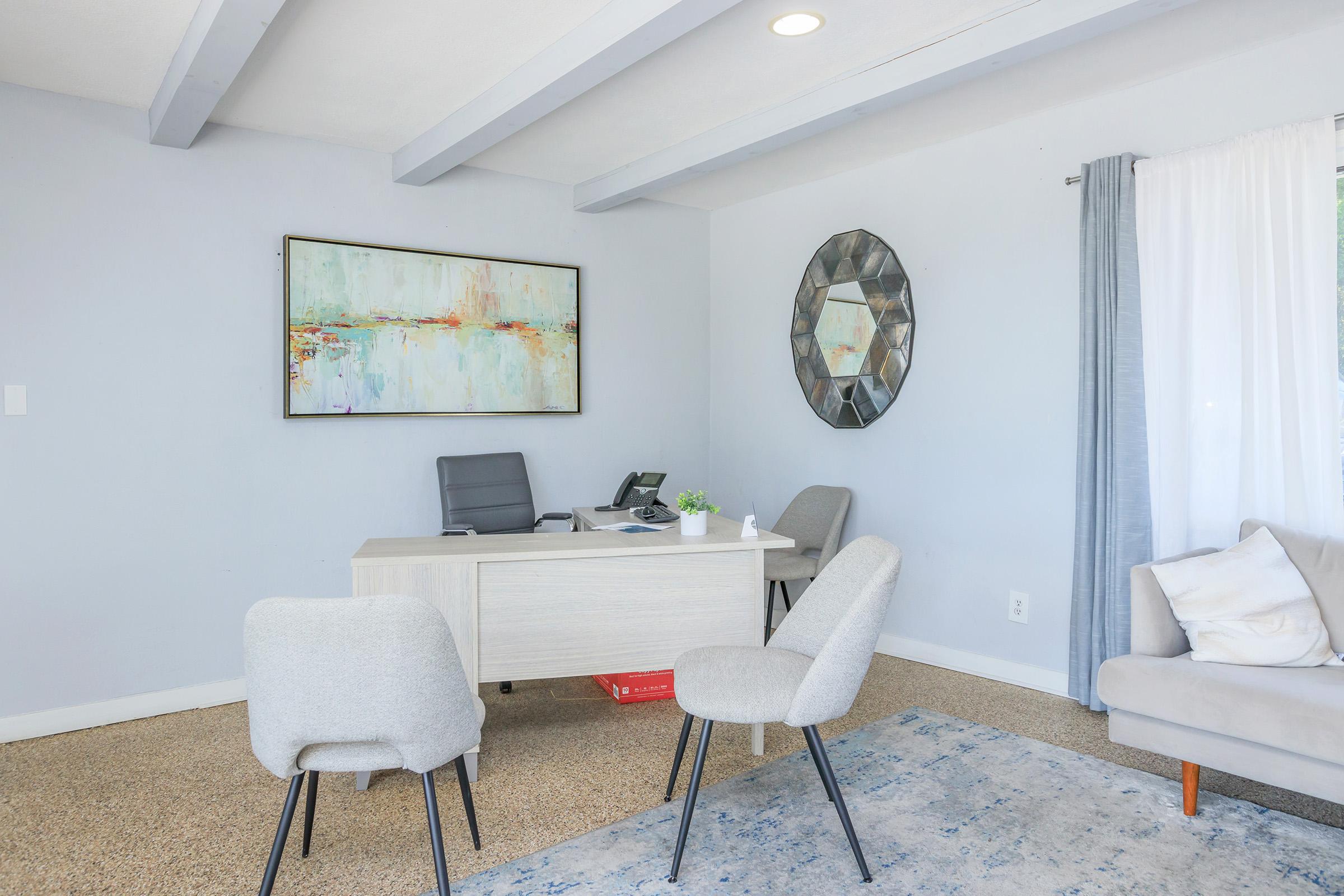 A modern office space featuring a pale blue wall, a stylish desk with a chair, and a telephone. There is a decorative mirror on the wall, an abstract painting above the desk, and light gray upholstered chairs. Natural light filters in through a window with sheer curtains, adding a welcoming ambiance.