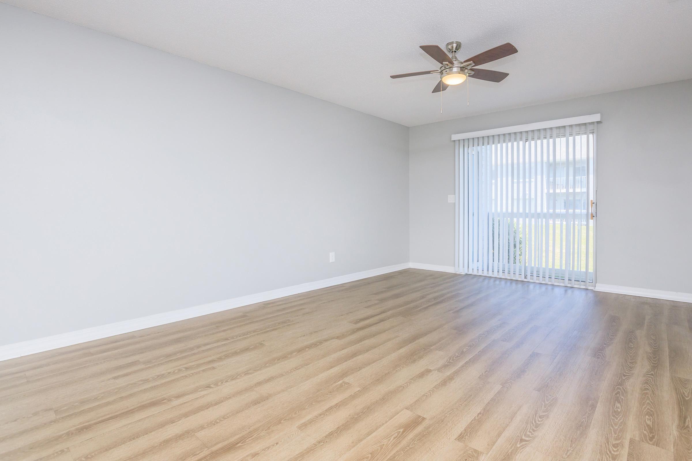 Spacious room with light gray walls and light wood flooring, featuring a ceiling fan. A large window with white vertical blinds allows natural light to enter. The room is empty, providing a blank canvas for decor and furnishings.