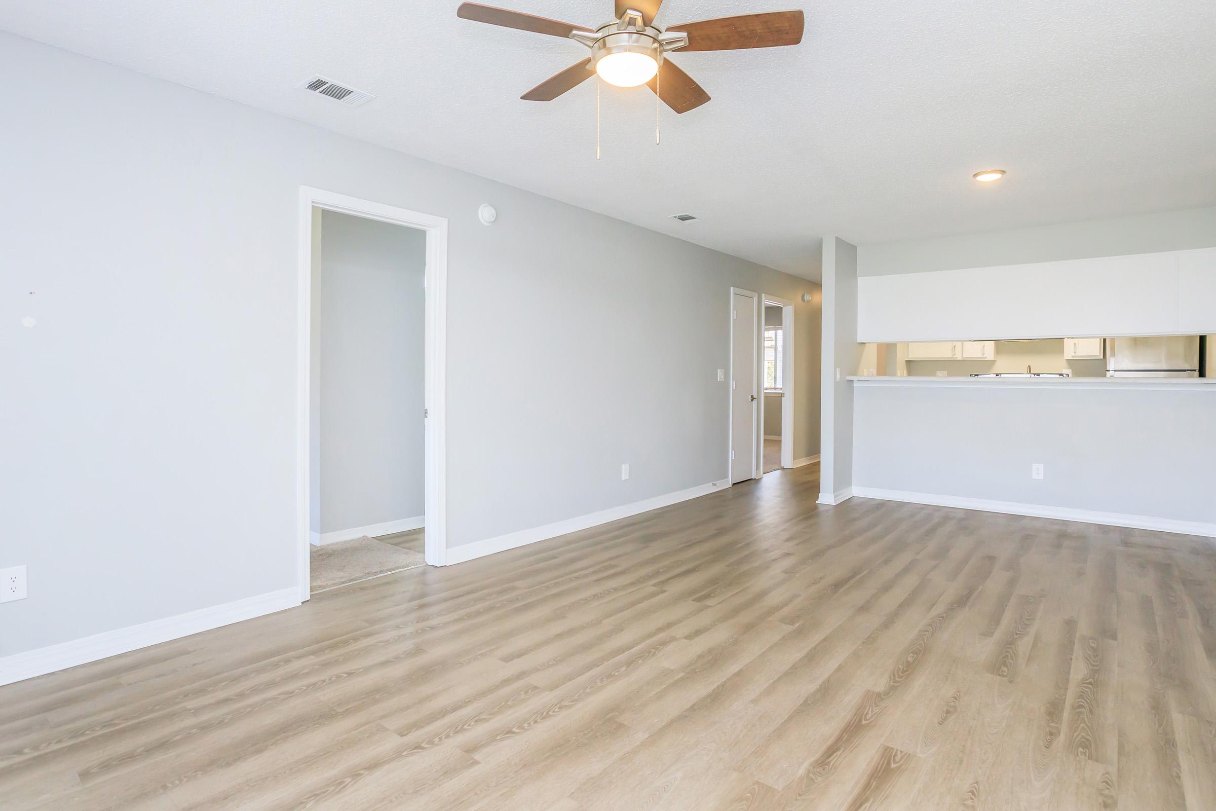 A spacious, well-lit room with light gray walls and modern wood flooring. A ceiling fan is mounted in the center. A doorway leads to another room on the left, while an opening on the right leads to a kitchen area. The space appears clean and ready for furnishing.