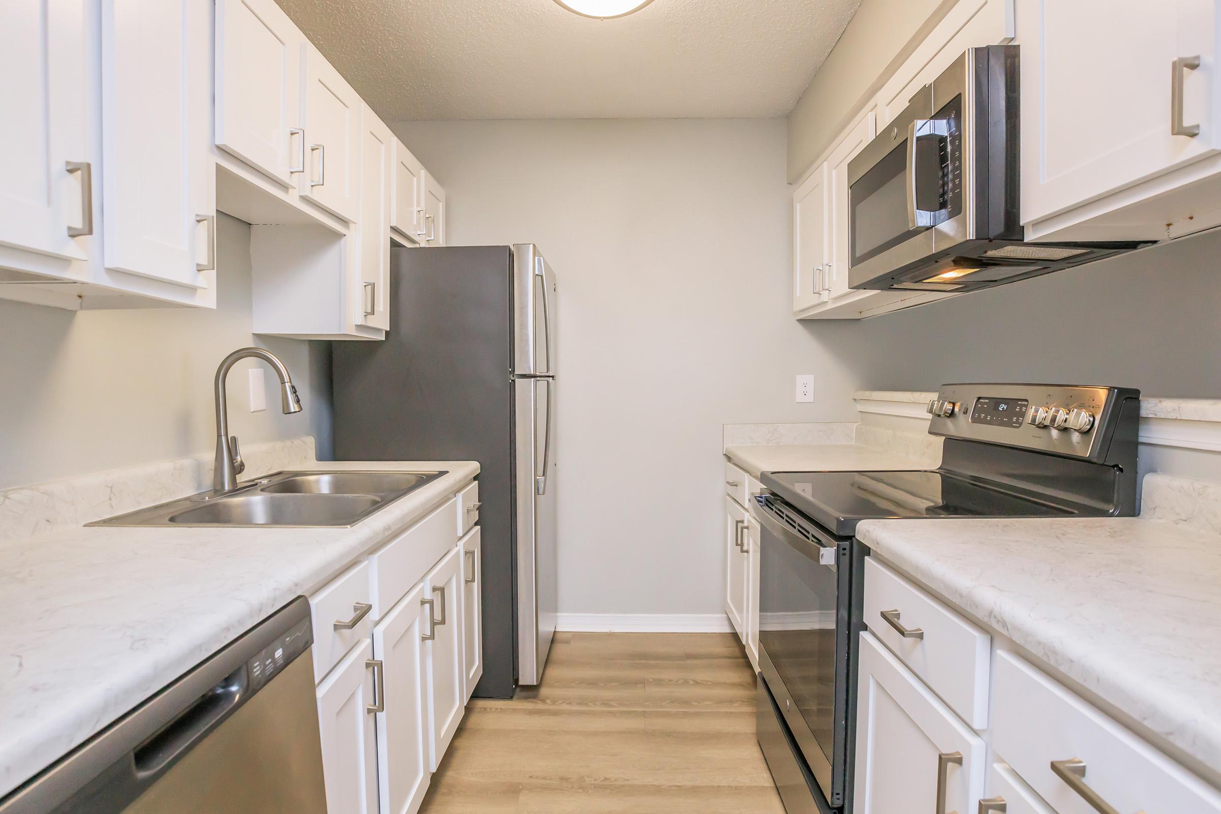 A modern kitchen featuring white cabinetry, a stainless steel refrigerator, an oven, and a microwave. It has a double sink with a sleek faucet and light-colored countertops. The walls are painted gray, and the flooring is wooden. Bright overhead lighting enhances the space.