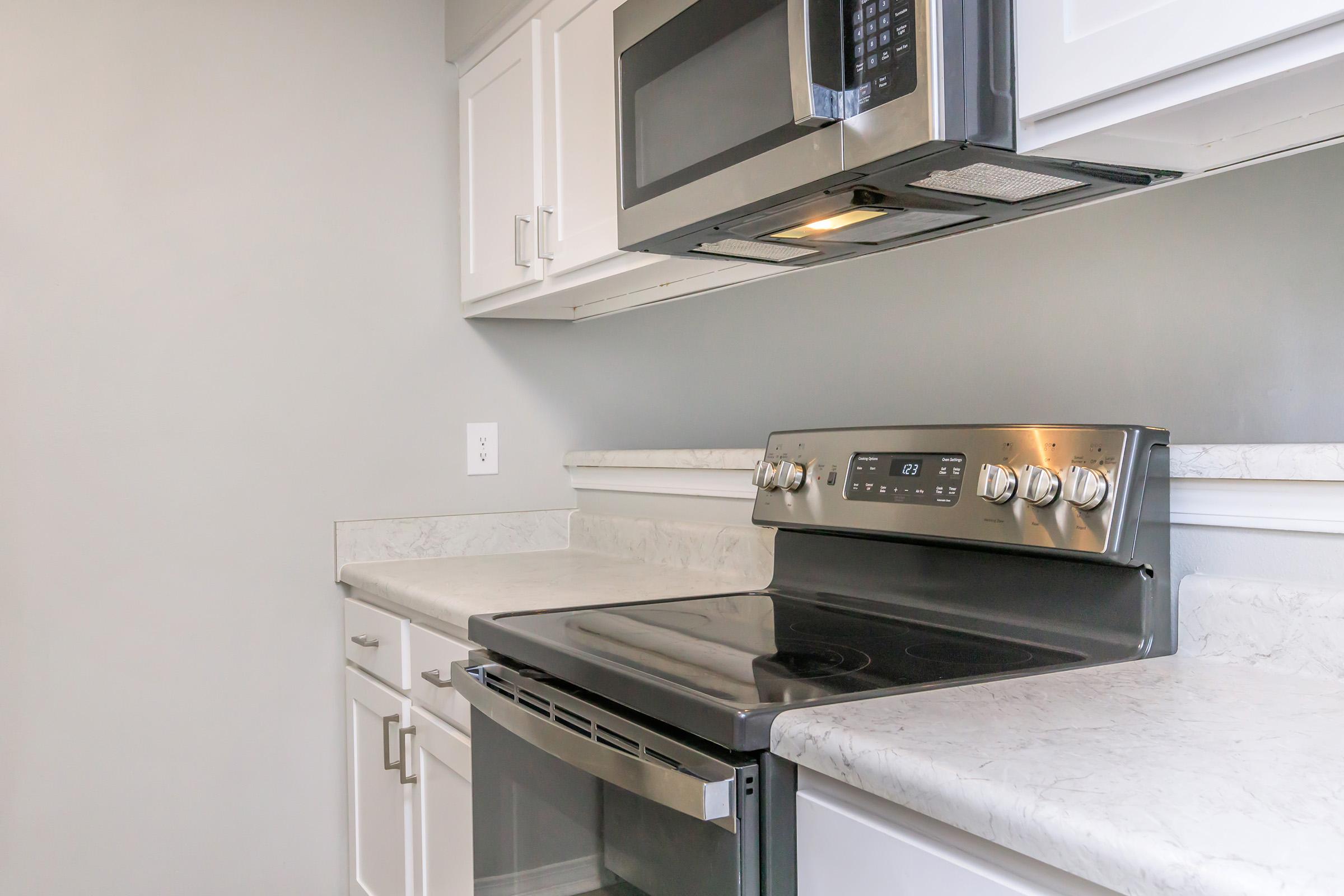 A modern kitchen featuring a stainless steel oven and a microwave above it. The countertops are made of light-colored stone, and the cabinetry is white, creating a clean and contemporary look. The walls are painted in a soft gray, contributing to a neutral and inviting ambiance.