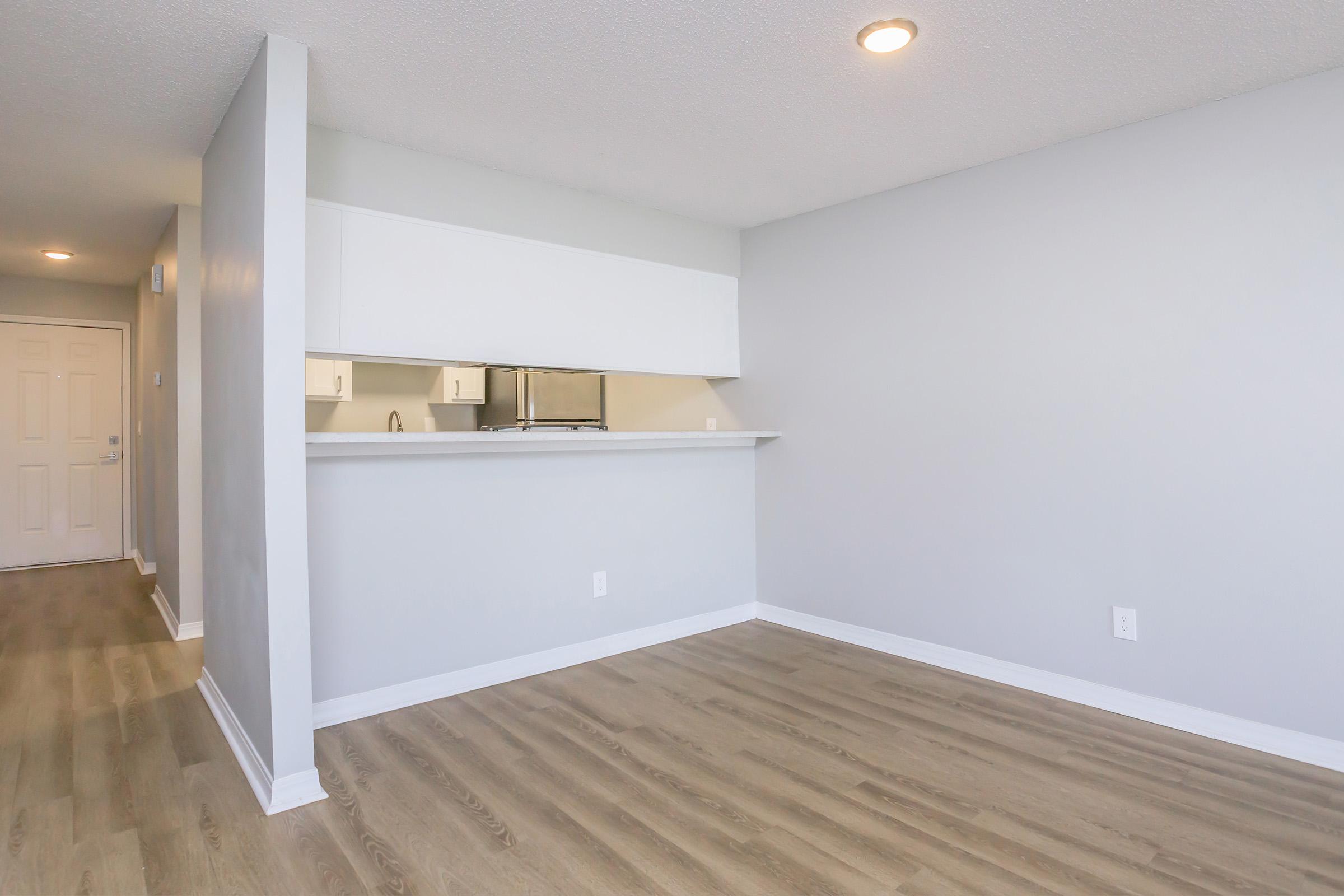 A spacious, modern living area featuring light-colored walls and wood laminate flooring. The room includes an open kitchen area with a counter bar, while a white door is visible on the left. The space is well-lit with overhead lighting and appears clean and uncluttered.