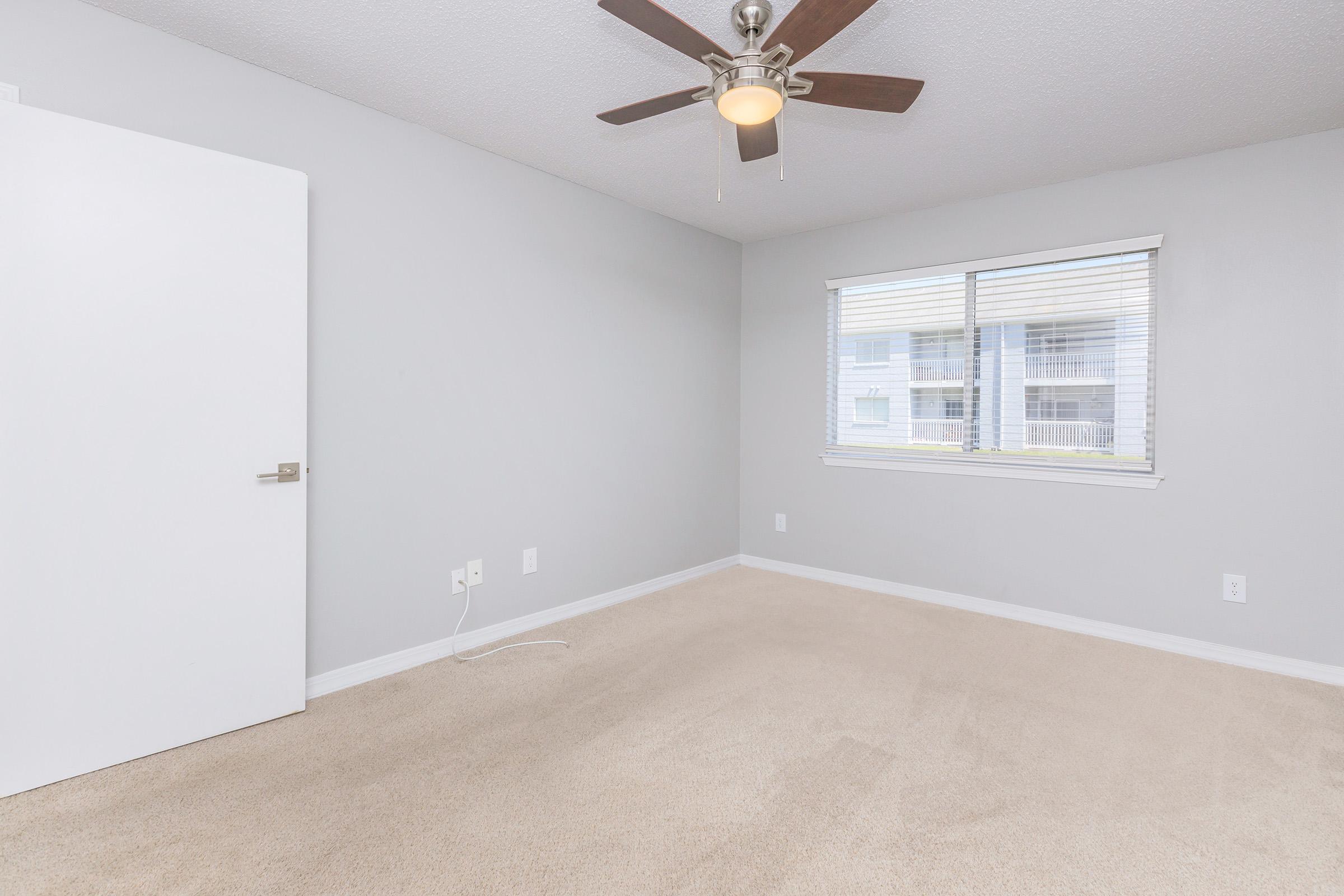 Empty room featuring light gray walls and carpet, a ceiling fan with wooden blades, and a white door. There is a large window with blinds allowing natural light, and the room has minimal furnishings for a clean, spacious appearance.