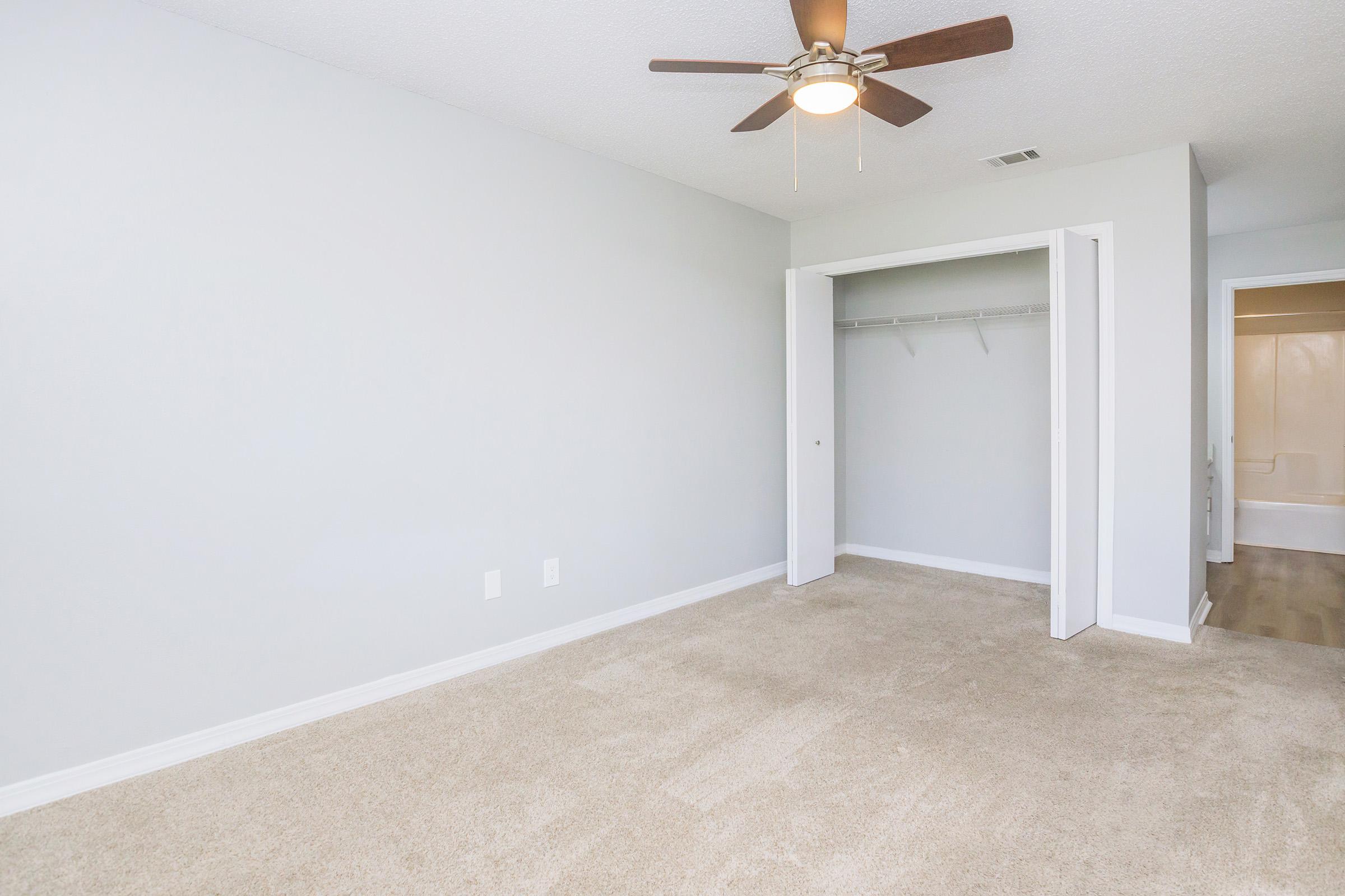 An empty room with light gray walls and beige carpet, featuring a ceiling fan. There's a closet with sliding doors on one side, and a bathroom entrance visible in the background. The space is well-lit, giving a bright and airy feel.