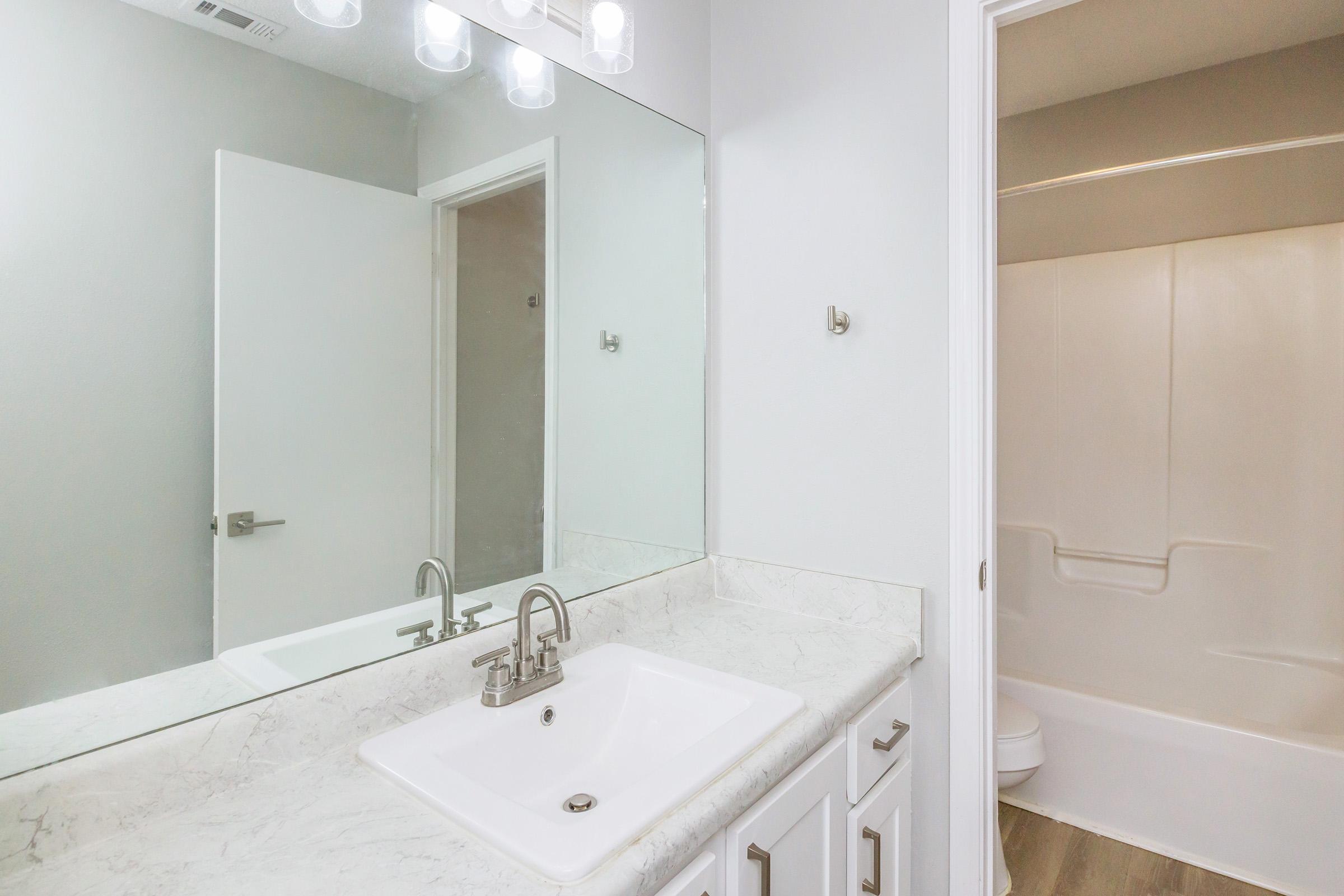 A modern bathroom featuring a white countertop with a sink, a large mirror above, sleek cabinetry, and a neutral color scheme. A shower/bath combo is visible in the background, along with a partially open door leading to another room. The space is well-lit with overhead lighting.