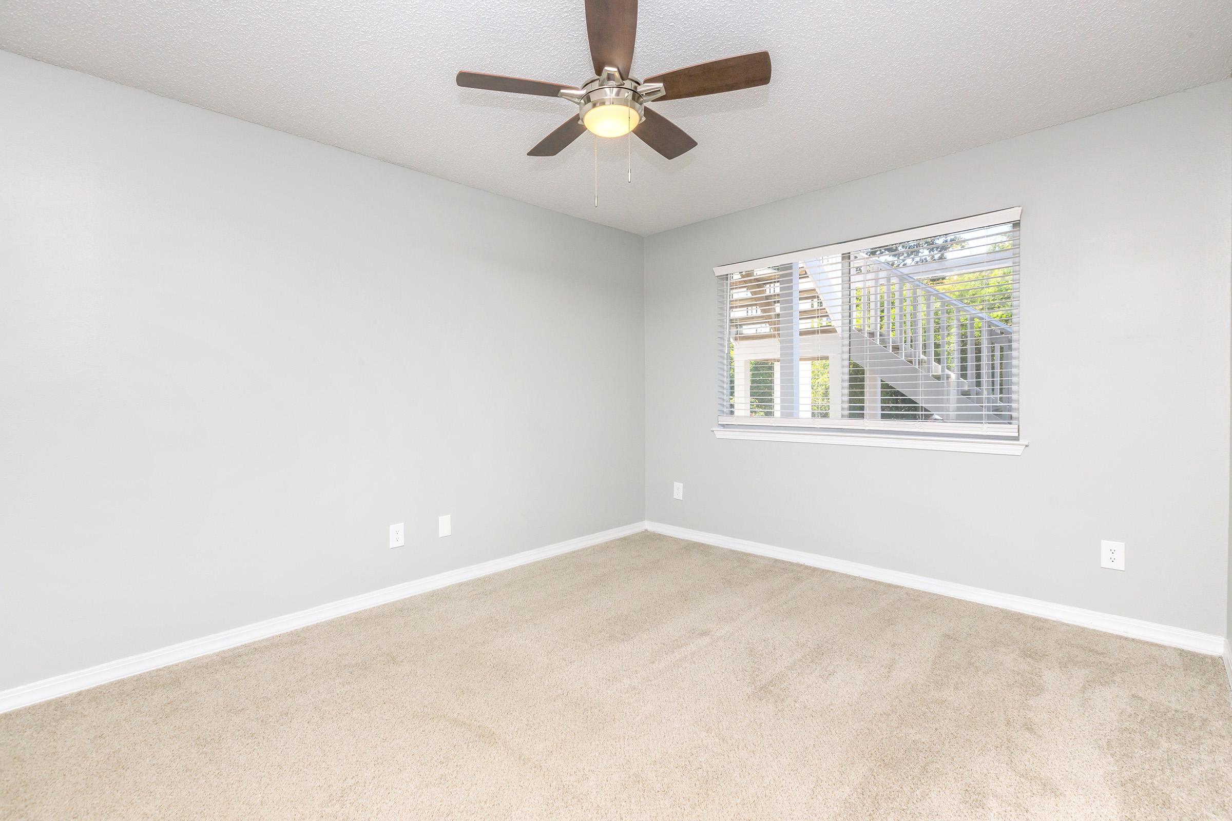 A well-lit, empty room with light gray walls and beige carpet. A ceiling fan is mounted above, and there's a window with horizontal blinds allowing natural light to enter. Outside the window, a glimpse of stairs leading to another level can be seen. The overall atmosphere is clean and spacious.