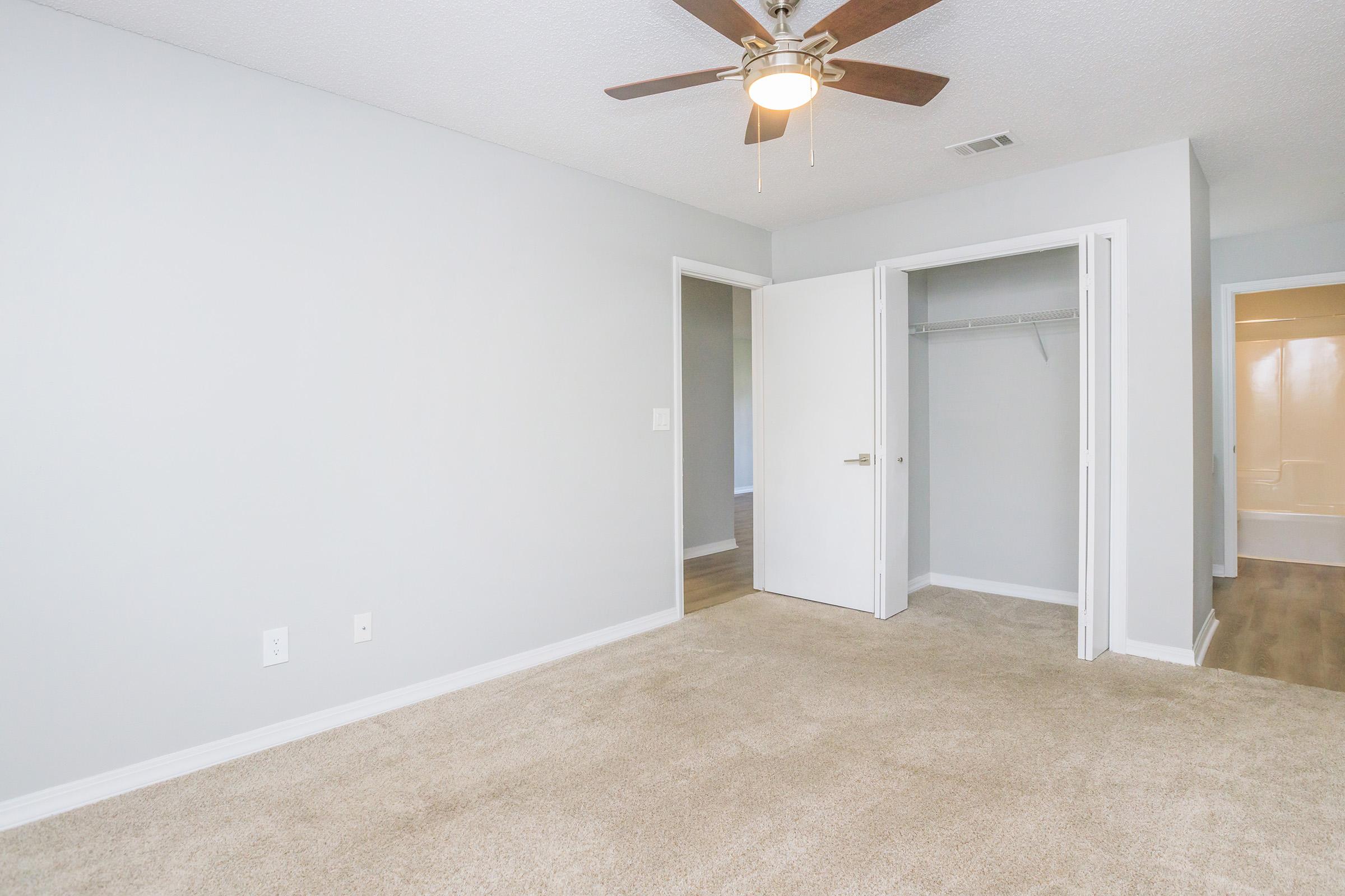 Empty room with light gray walls and carpeted floor. A ceiling fan is visible, along with an open closet and two doors leading to other spaces. The lighting is bright, highlighting the room's spaciousness and clean lines.