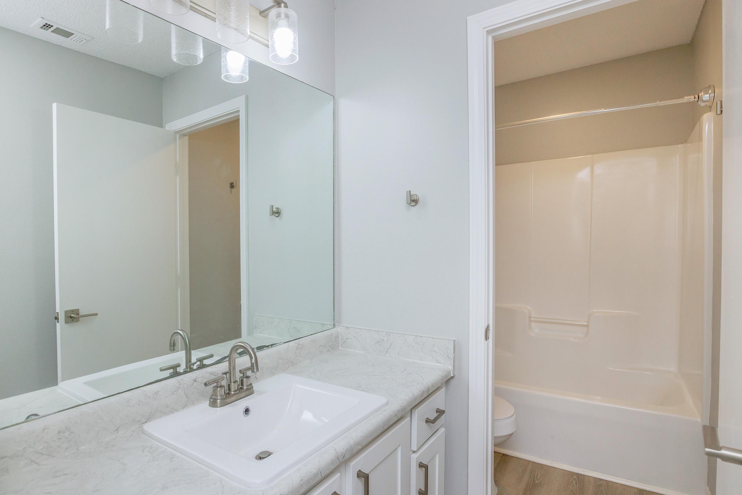 A modern bathroom featuring a marble countertop sink with a chrome faucet, a large mirror above the sink, and light fixtures. To the right, there is a separate area with a bathtub and shower combination, and a toilet in a neutral-colored space with light gray walls.