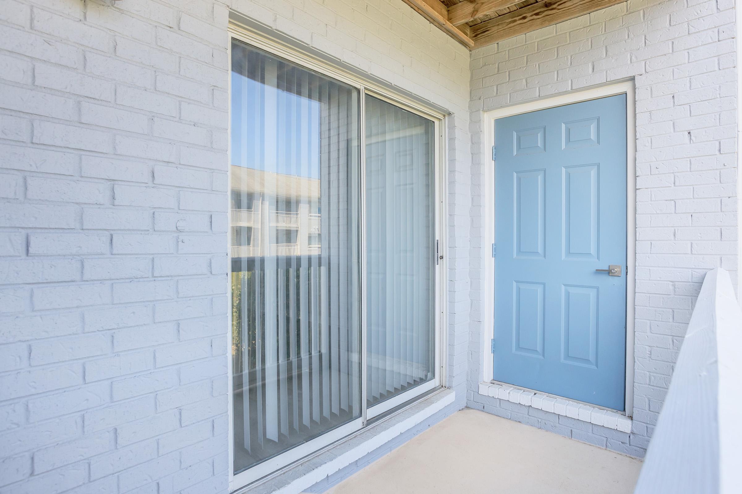 A simple balcony with a sliding glass door and a blue door. The wall is painted white brick, and there are sheer curtains visible through the glass. The balcony has a concrete floor and a wooden overhead structure, suggesting an outdoor living space.